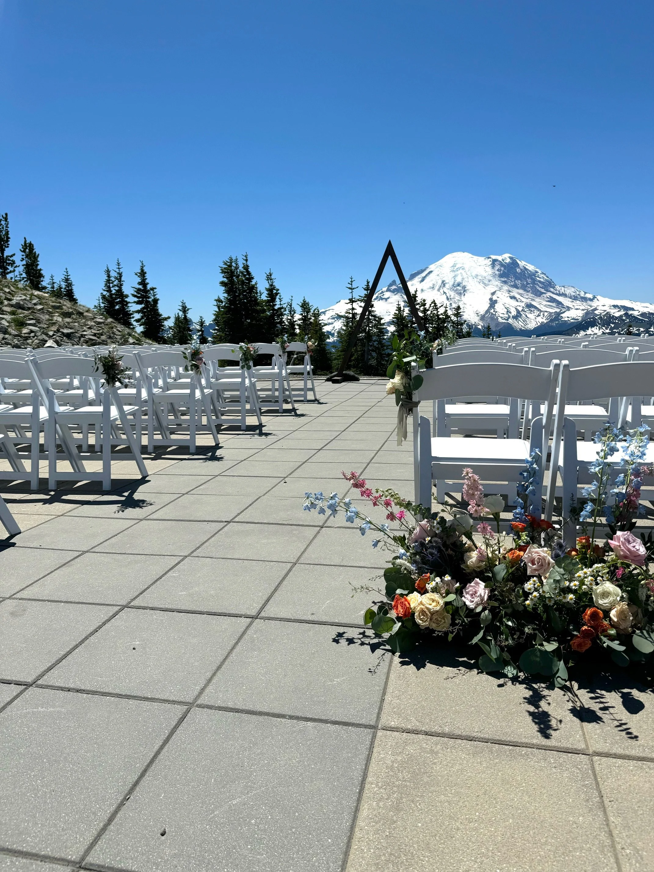 Outdoor wedding setup with white chairs decorated with flowers, an altar with a mountain and snow-covered peak in the background, and a large flower arrangement on the ground.