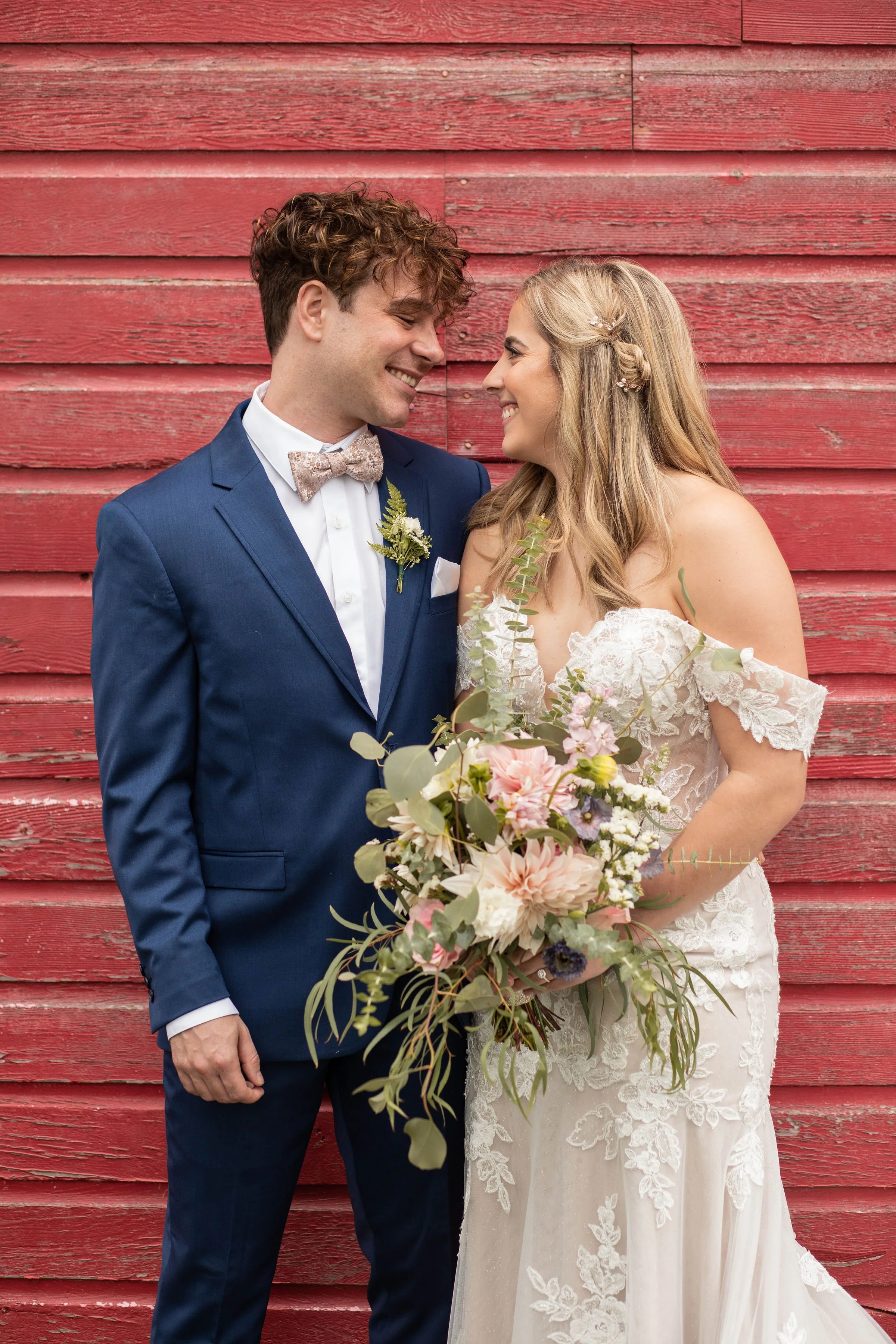 A bride and groom stand close together, smiling at each other, in front of a red wooden wall. The bride is holding a large bouquet of flowers and wearing an off-shoulder lace wedding dress. The groom is in a blue suit with a patterned bow tie.
