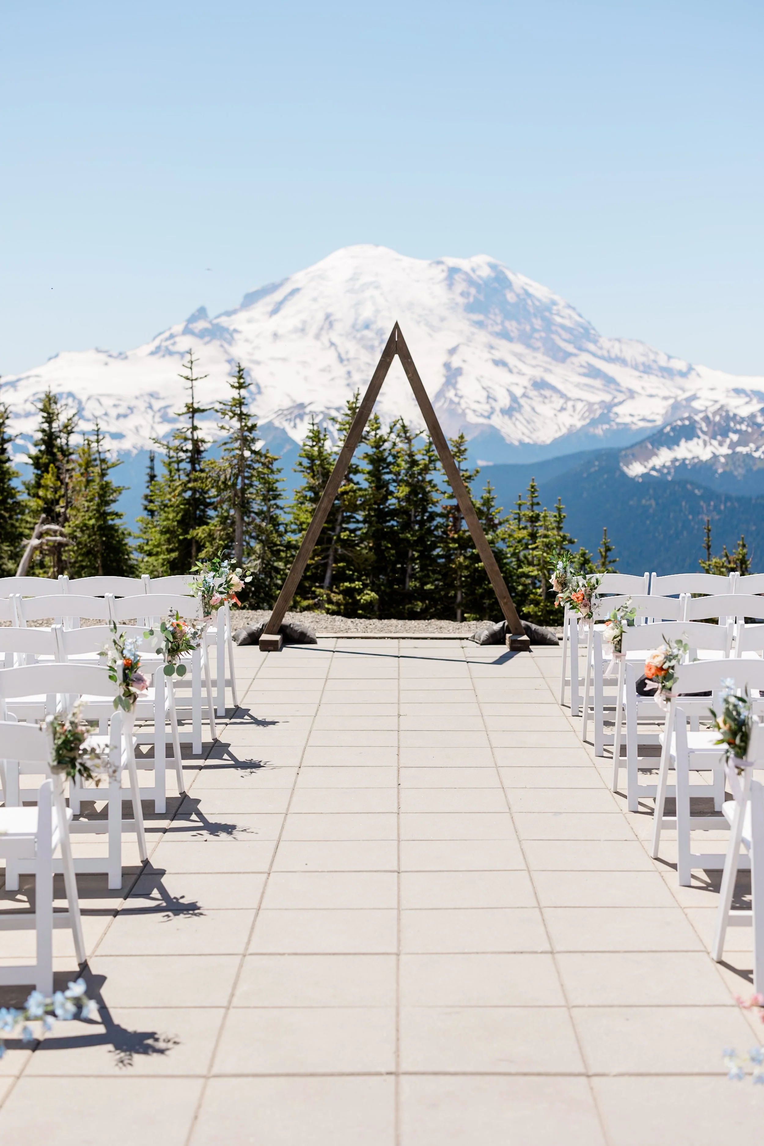 Outdoor wedding setup with white chairs decorated with flowers, a wooden arch, evergreen trees, and Mount Rainier in the background under a clear blue sky.