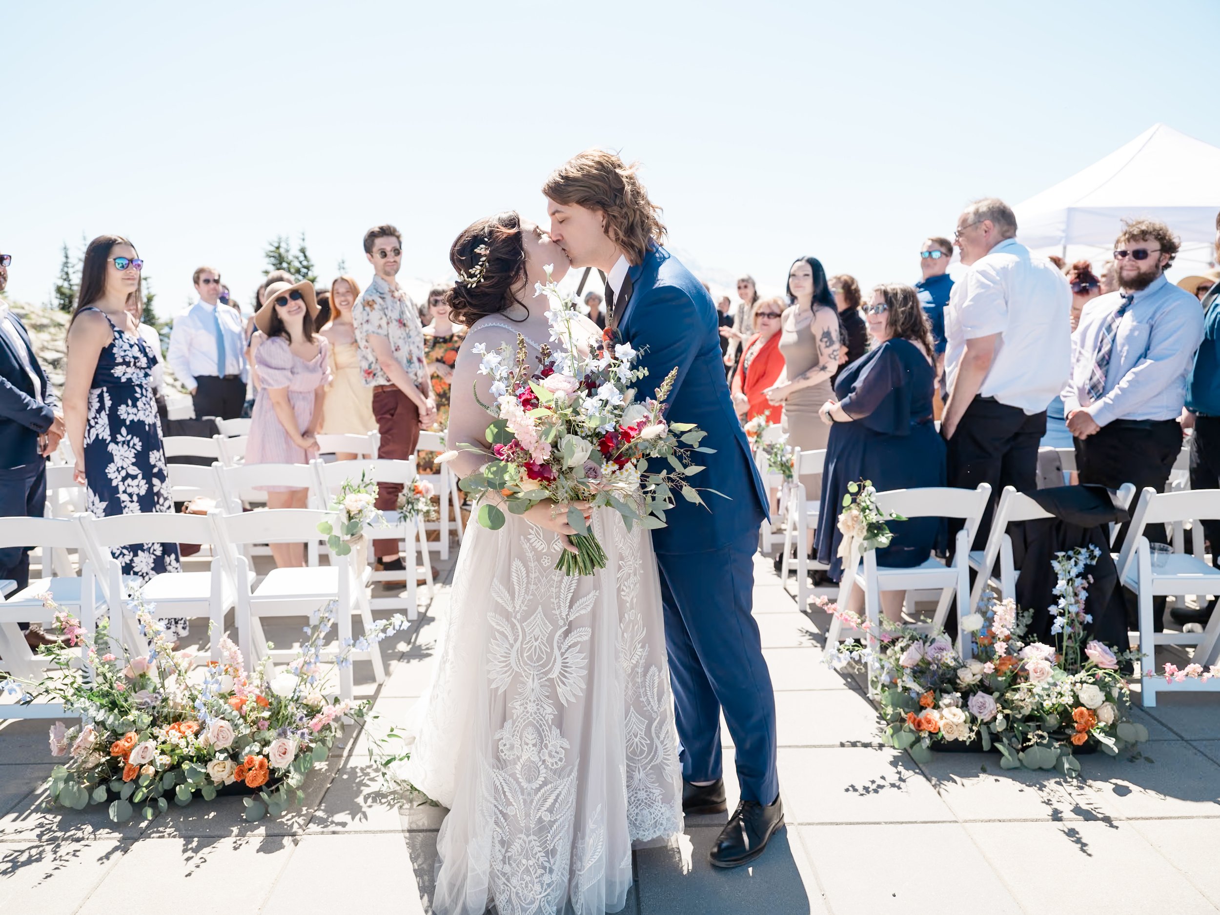A bride and groom kiss during an outdoor wedding ceremony on a sunny day, surrounded by guests and floral arrangements.