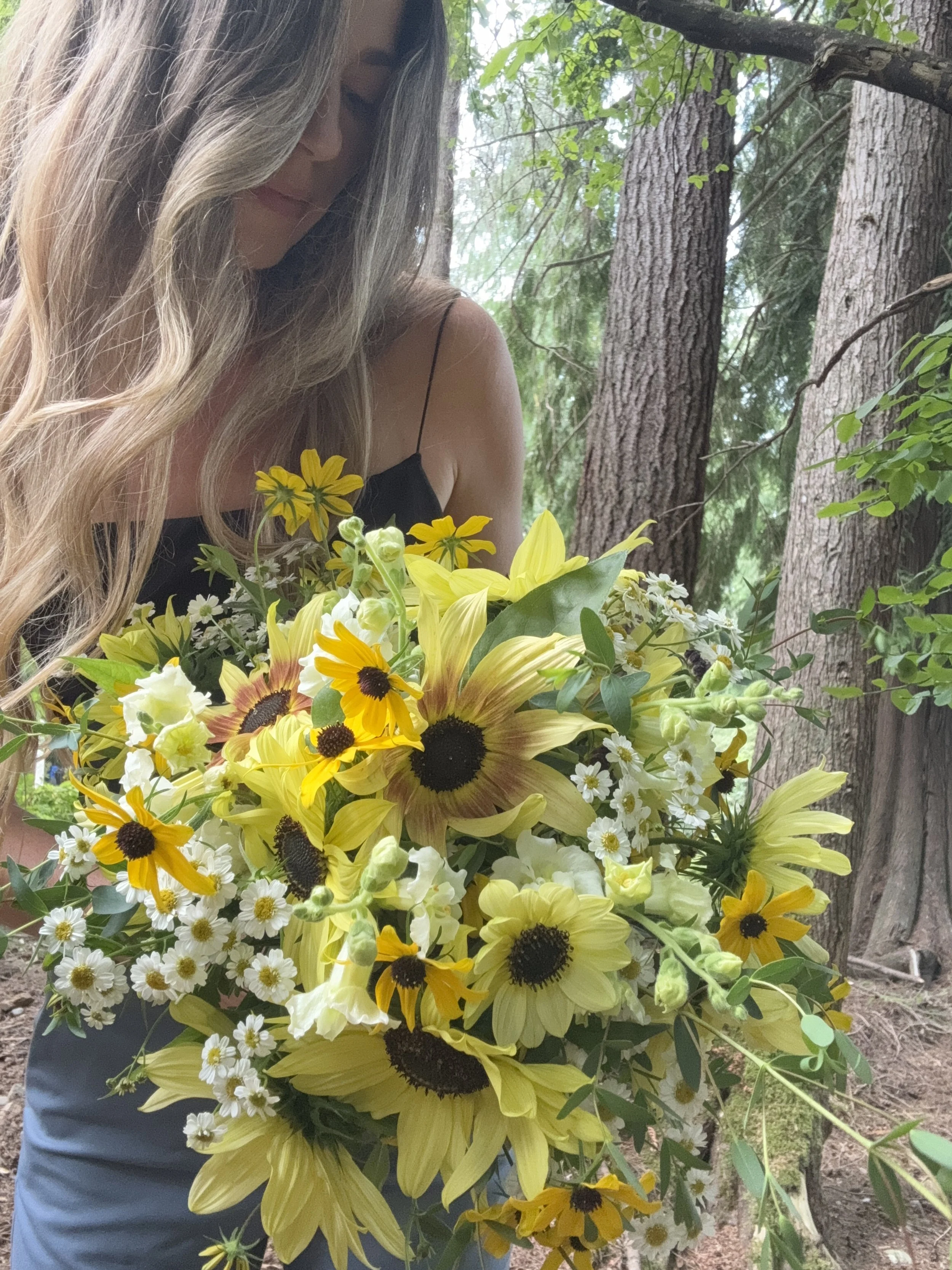 A woman with long, wavy blonde hair in a black spaghetti strap top holding a large bouquet of yellow and white flowers, with trees in the background.