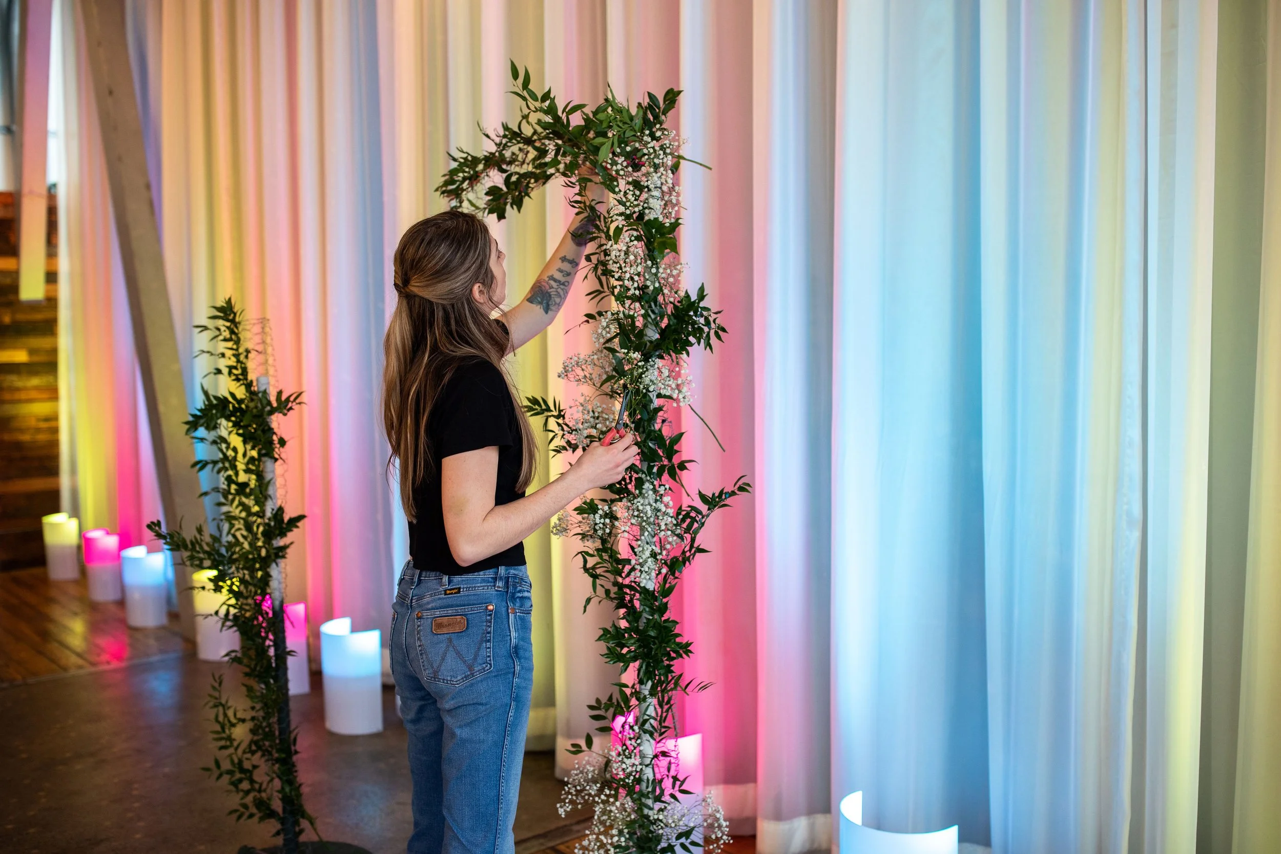 A woman with long brown hair and tattoos on her arm decorating with flowers at a colorful event space. She is wearing a black t-shirt and jeans, arranging a flower arrangement against a backdrop of pastel-colored curtains with lighting.