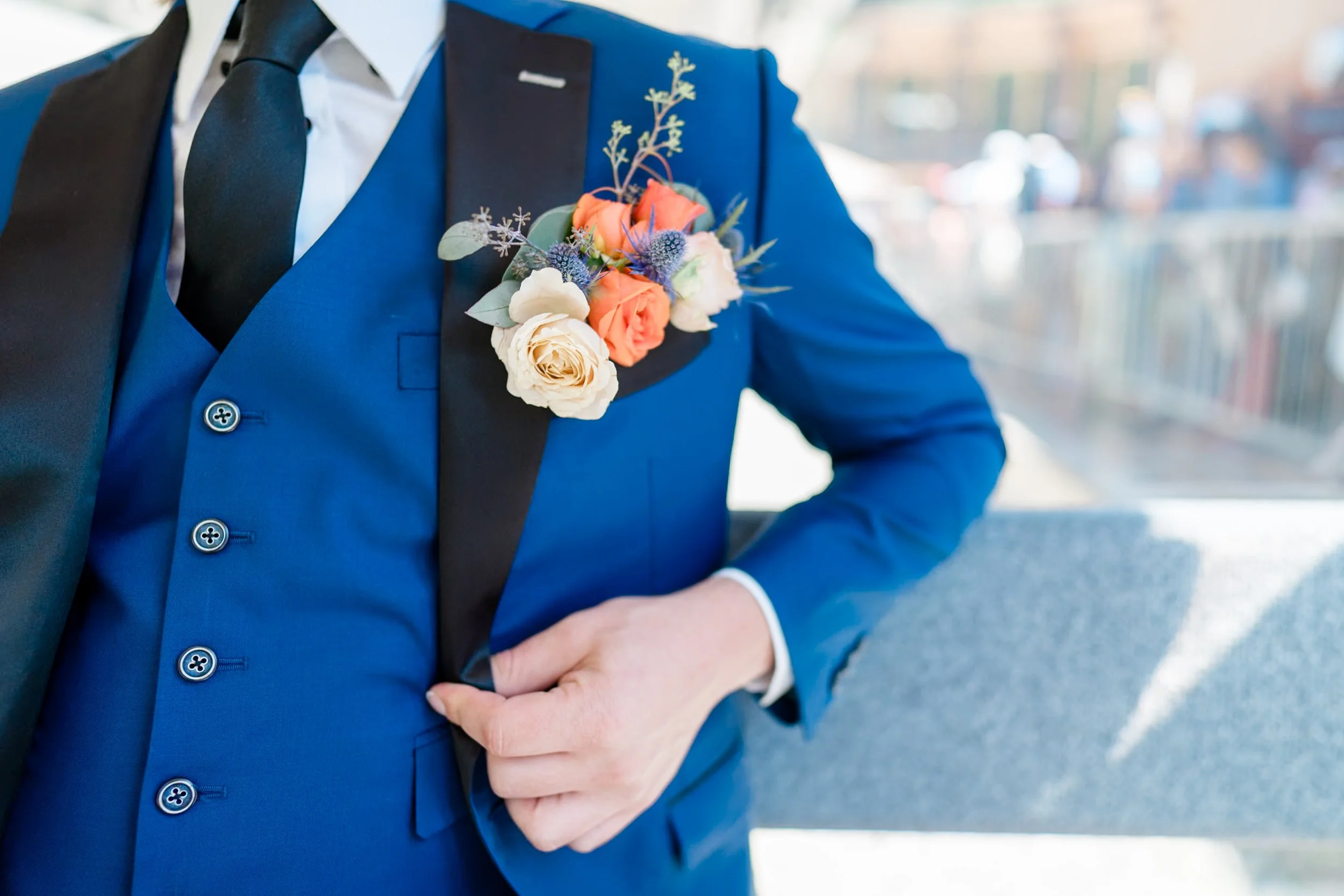 Close-up of a man in a blue tuxedo with a black lapel, white shirt, and black tie, wearing a colorful boutonniere on his lapel, standing outdoors with a blurred city background.
