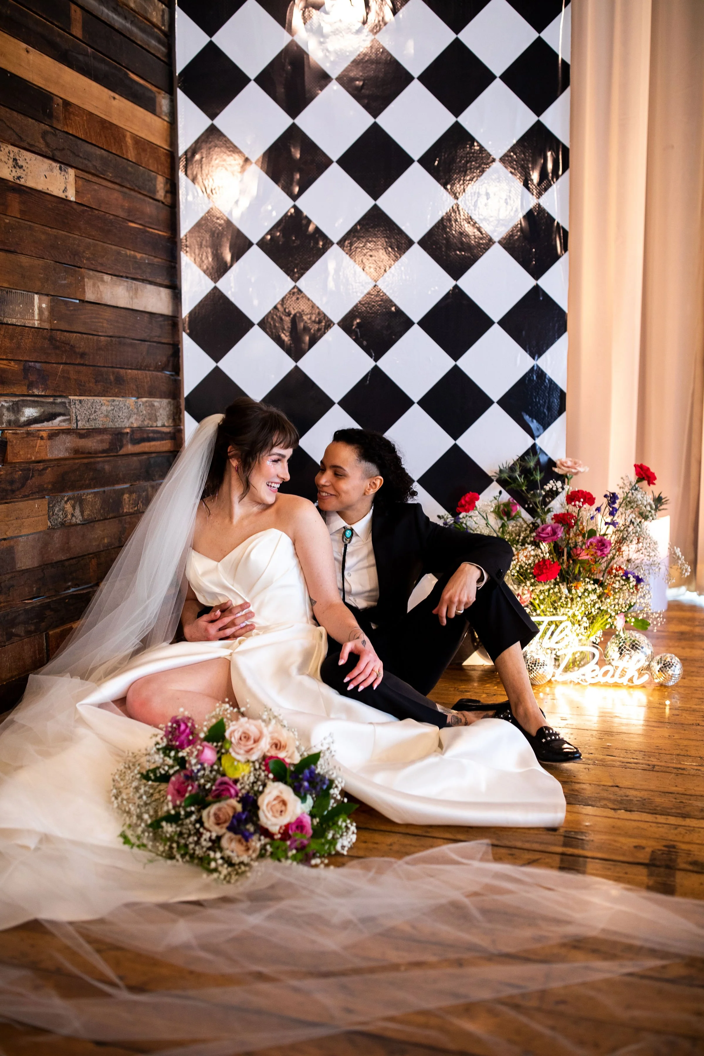 Two women sitting on a wooden floor with floral arrangements, one in a wedding dress and veil, the other in a black suit, smiling and looking at each other, indoor wedding celebration.