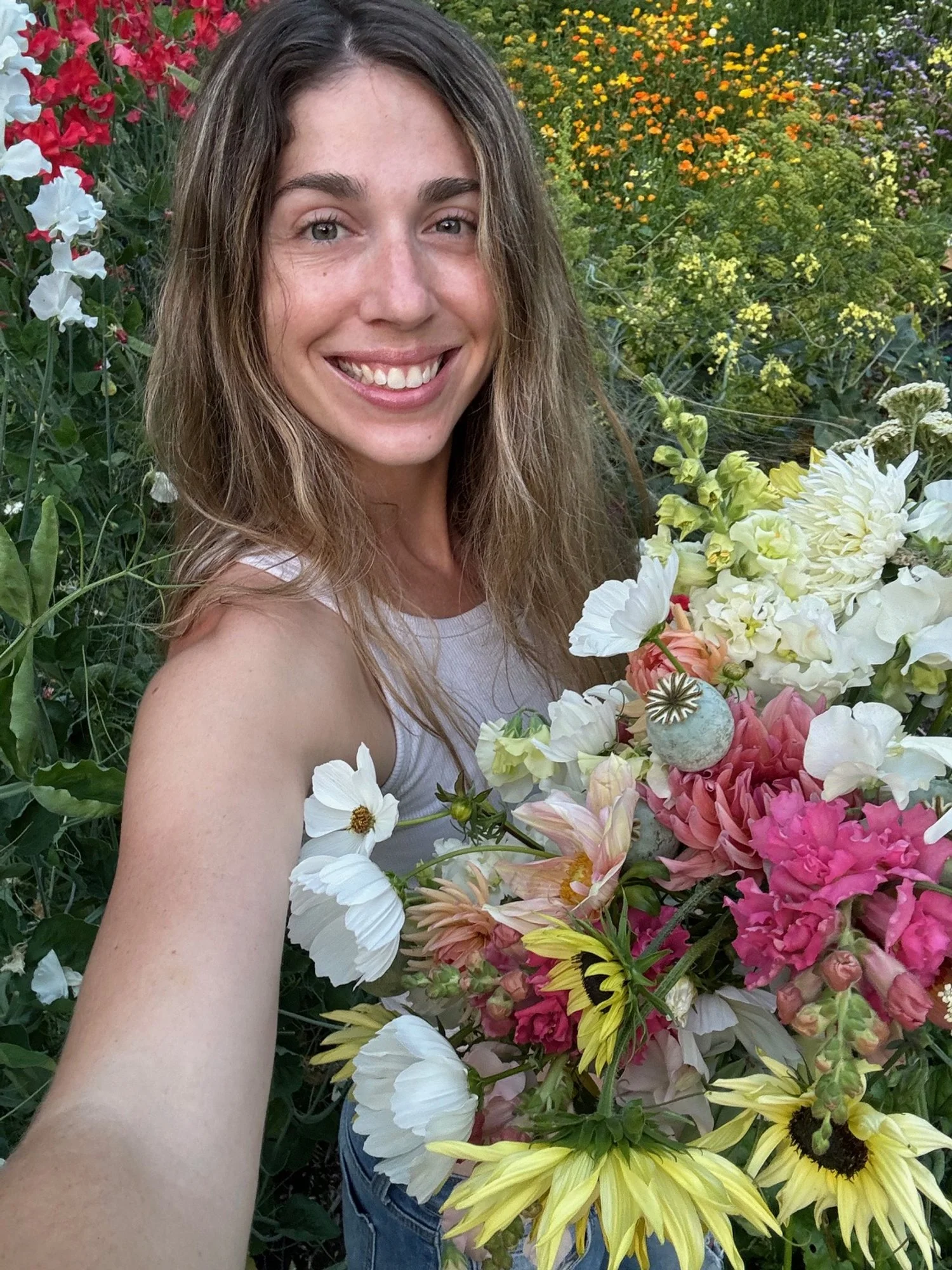 A woman taking a selfie in a garden with a large bouquet of various colorful flowers, including white, yellow, pink, and orange blooms, with a background of green foliage and additional flowers.