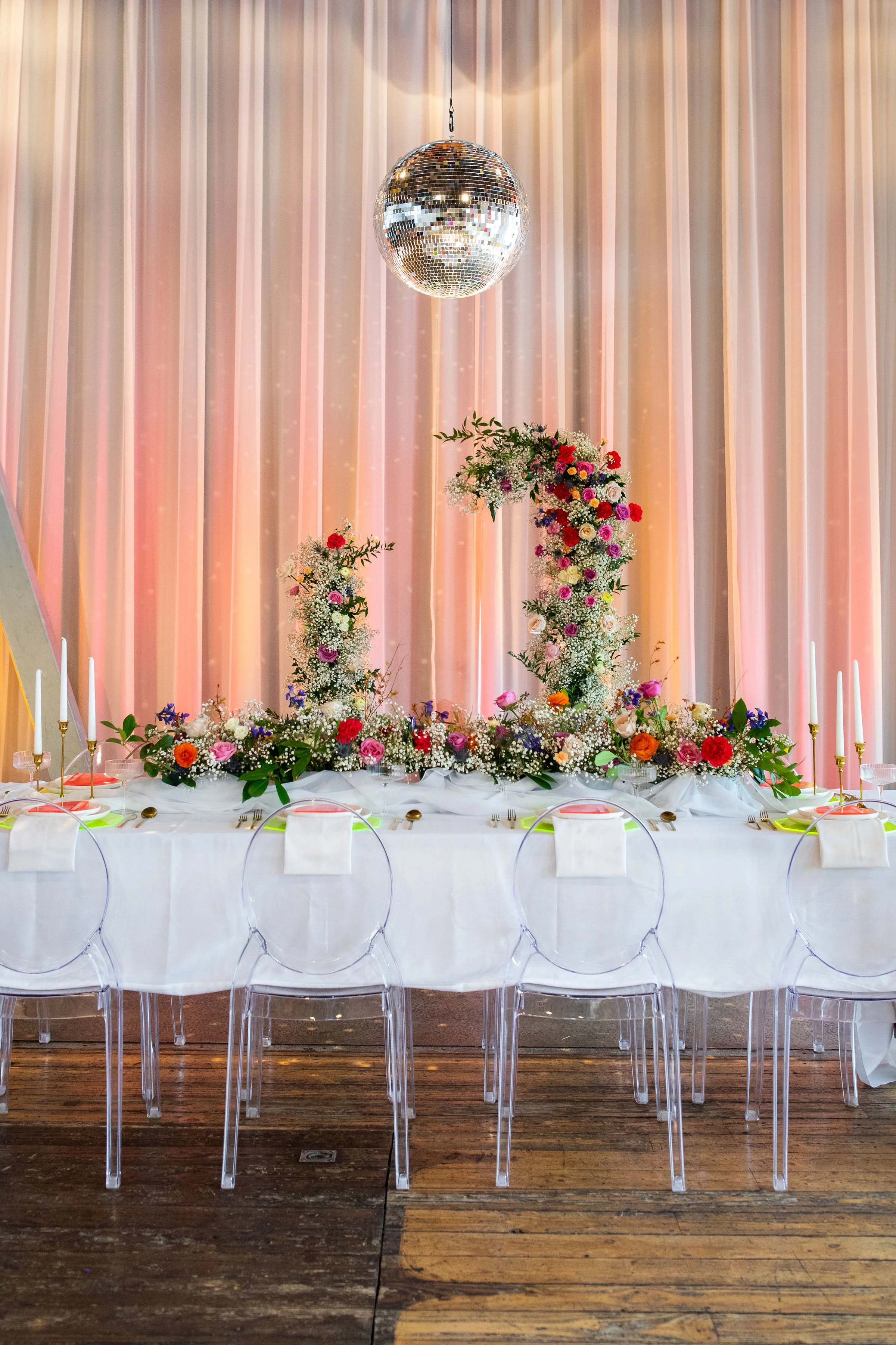 Elegant event table with flowers, candles, and a disco ball hanging from the ceiling, against a pink curtain backdrop.
