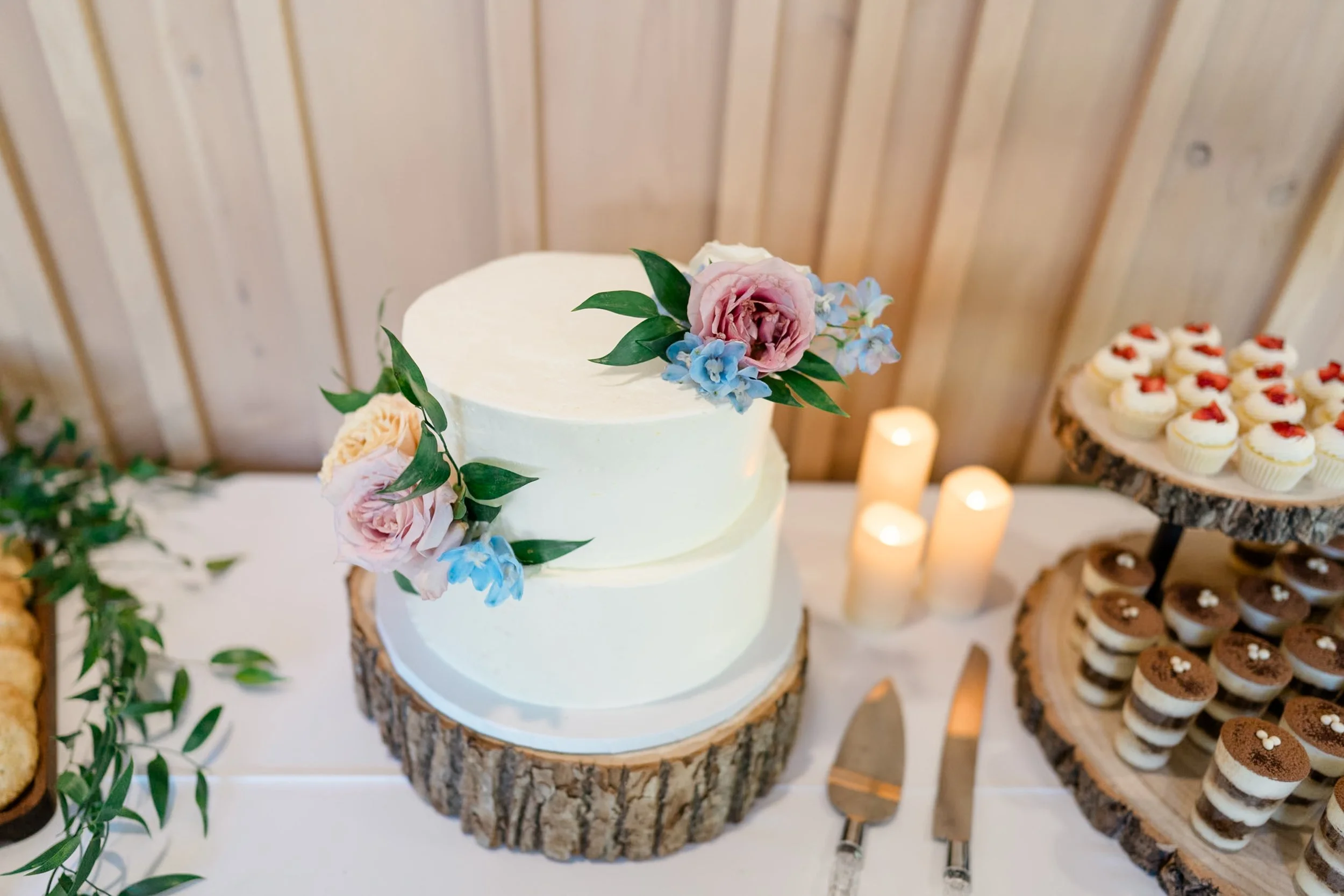 A decorated two-tier wedding cake with pink, blue, and white flowers on top. The cake sits on a wooden log slice. To the right, there are cupcakes with white frosting and red topping, along with some layered desserts in small glasses. Candle lights a
