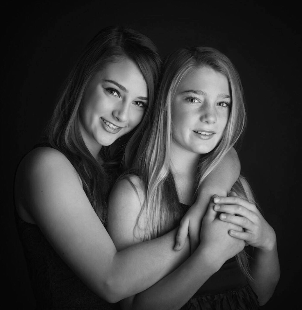 Two young women with long hair smiling and hugging each other in a black-and-white portrait against a dark background.