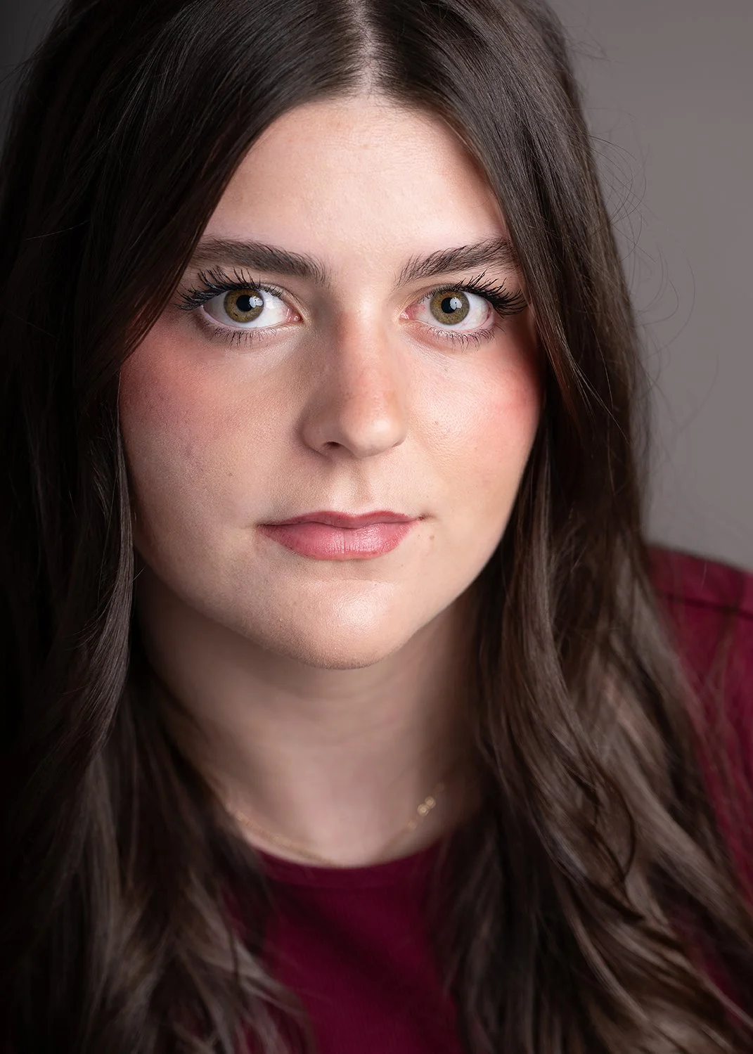 Close-up portrait of a young woman with long brown hair, green eyes, light makeup, and a neutral expression, wearing a burgundy top against a plain background.