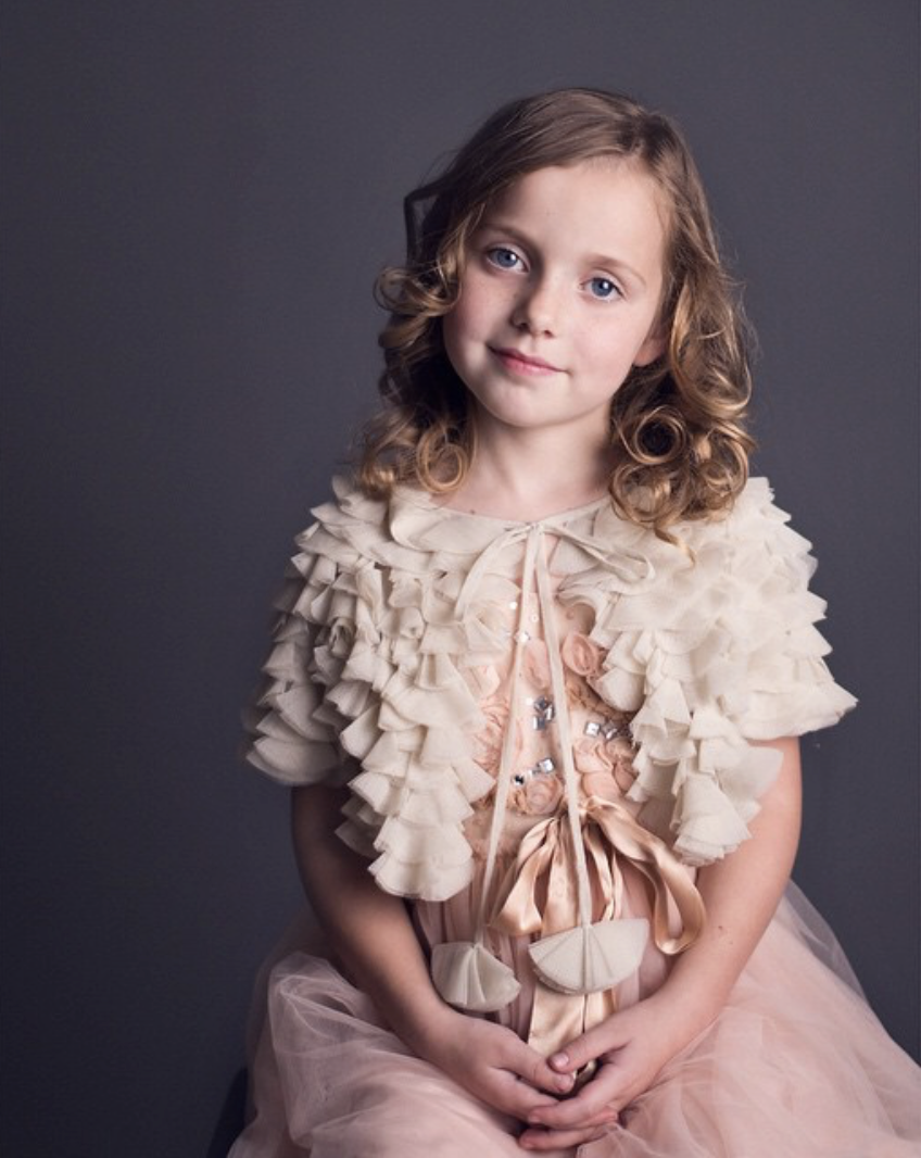 A young girl with curly blonde hair and blue eyes, wearing a light pink dress with ruffles and decorative elements, sitting against a dark gray background.