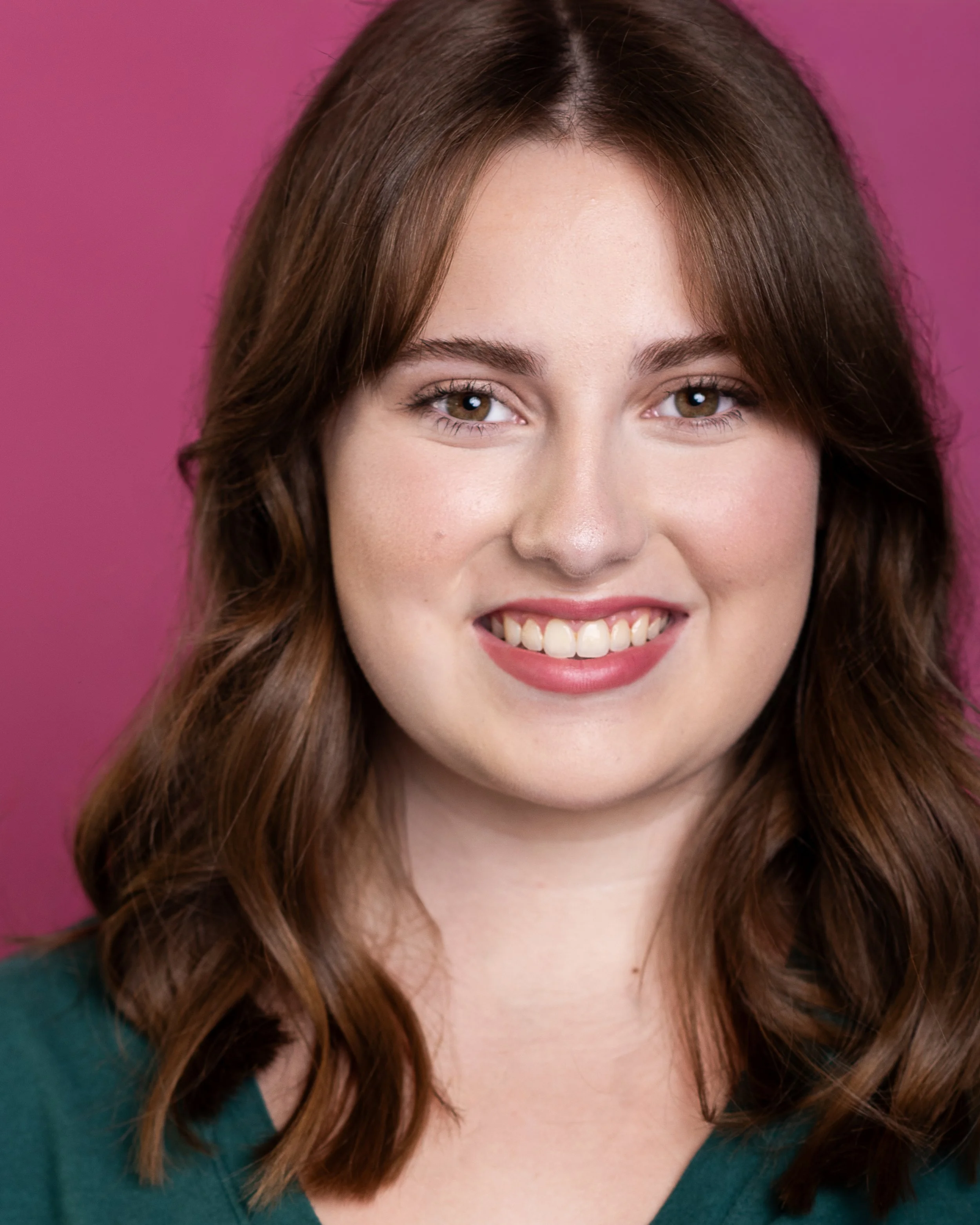 Portrait of a young woman with light brown hair, smiling, wearing a green top, against a pink background.