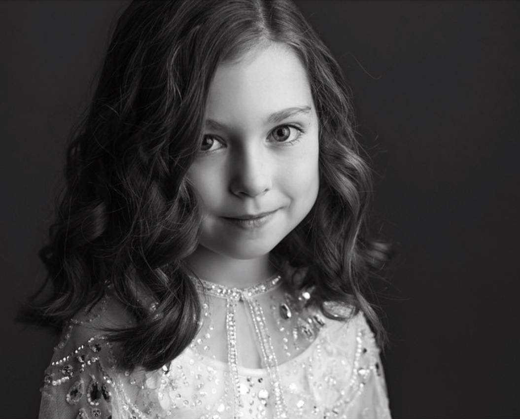 Black and white portrait of a young girl with shoulder-length wavy hair, looking at the camera with a slight smile, wearing a dress with intricate beading and embellishments.