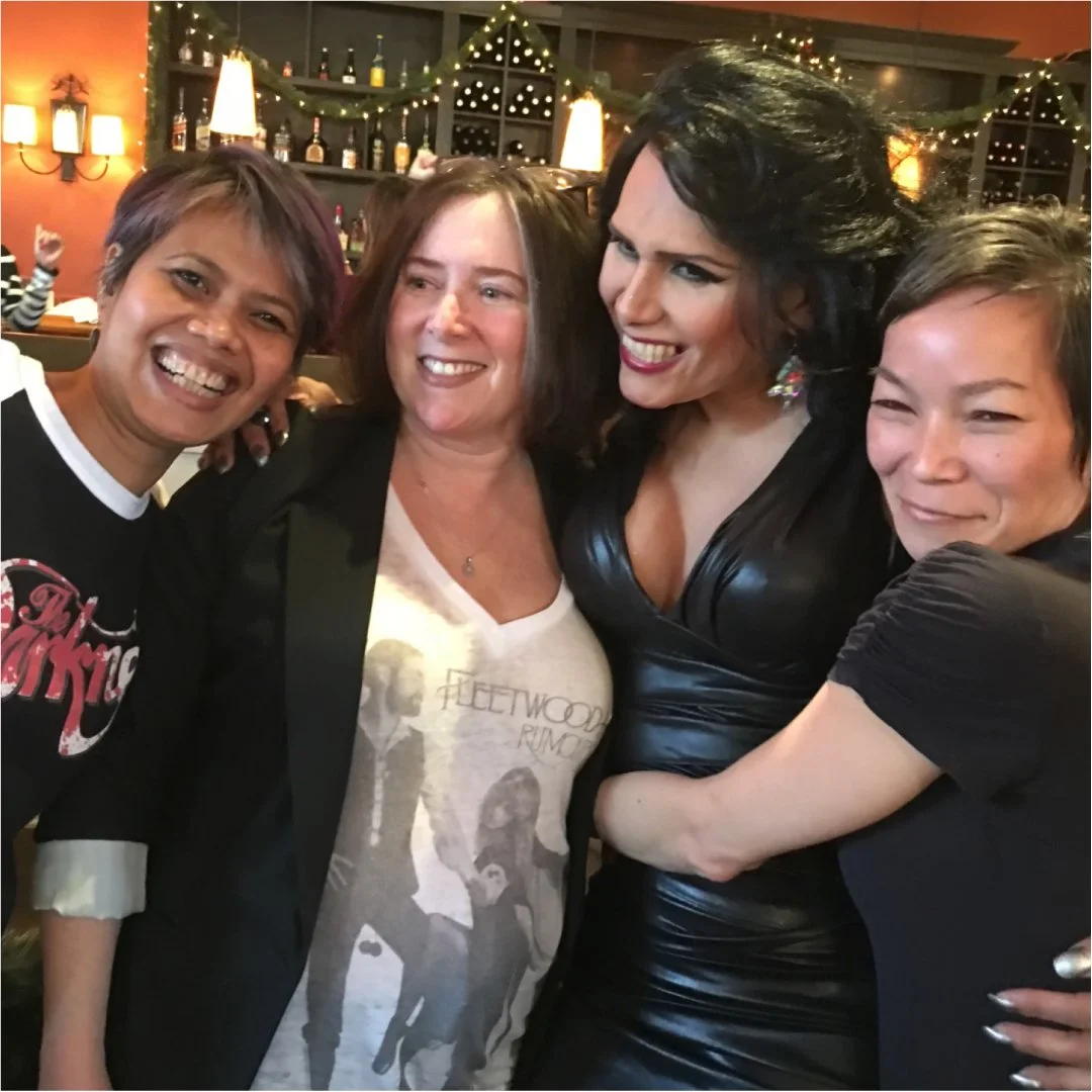 Four women smiling and posing closely together at a festive indoor gathering with Christmas lights and decorations in the background.