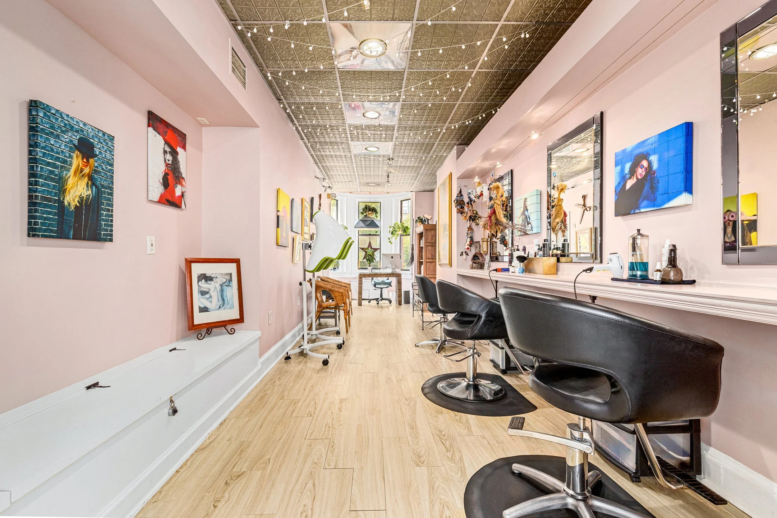Interior of an art studio or salon with light pink walls, various artwork on display, black salon chairs, and decorative items. Large window at the end bringing in natural light.
