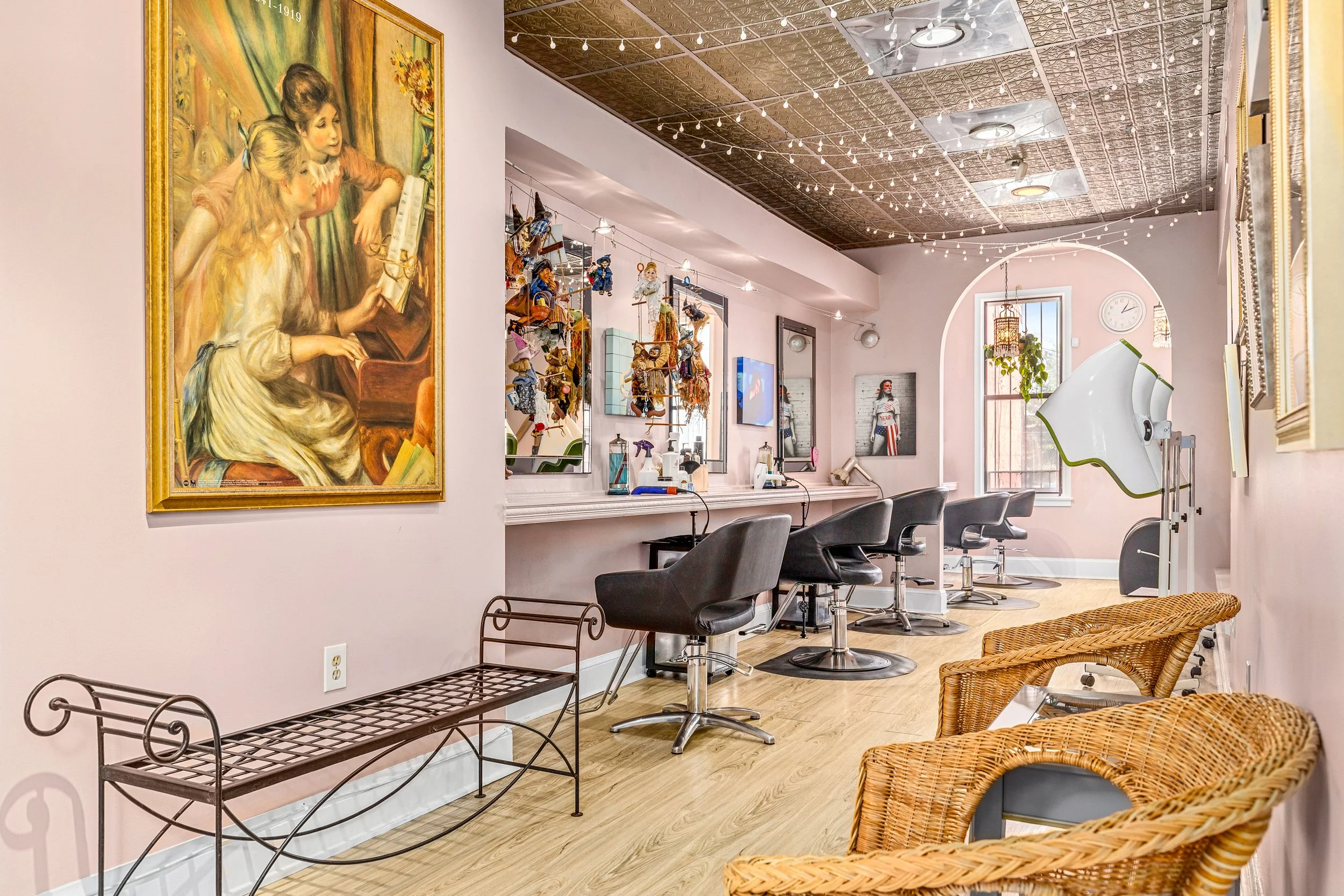 Interior of a hair salon with pink walls, black styling chairs, and a mirror wall. Artworks and decorations line the walls, with wicker chairs in the foreground and a styling station with hairdressing tools. String lights hang from the ceiling.