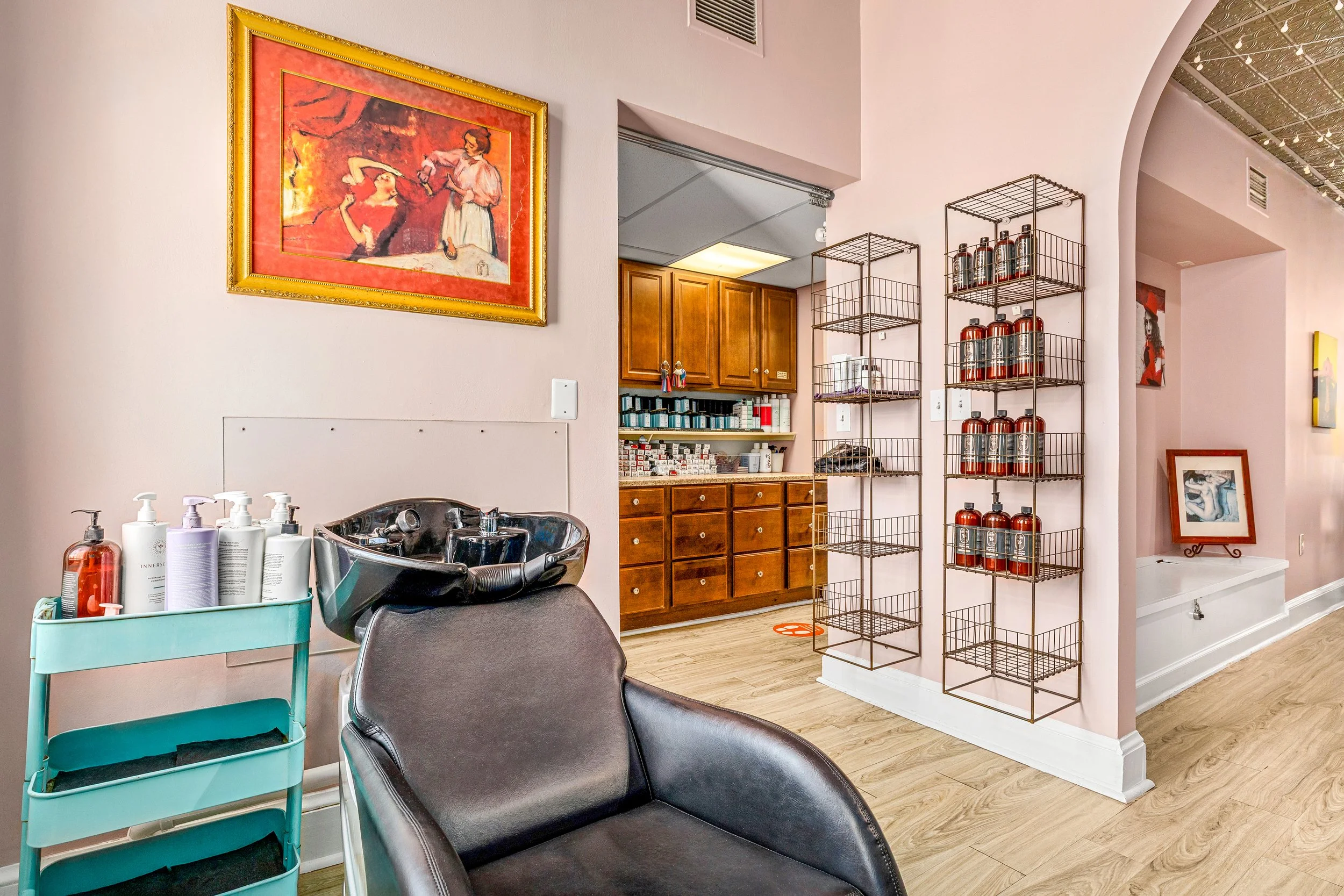 Interior of a hair salon or beauty spa with a styling chair, hair washing sink, shelves with hair care products, and artwork on pink walls.
