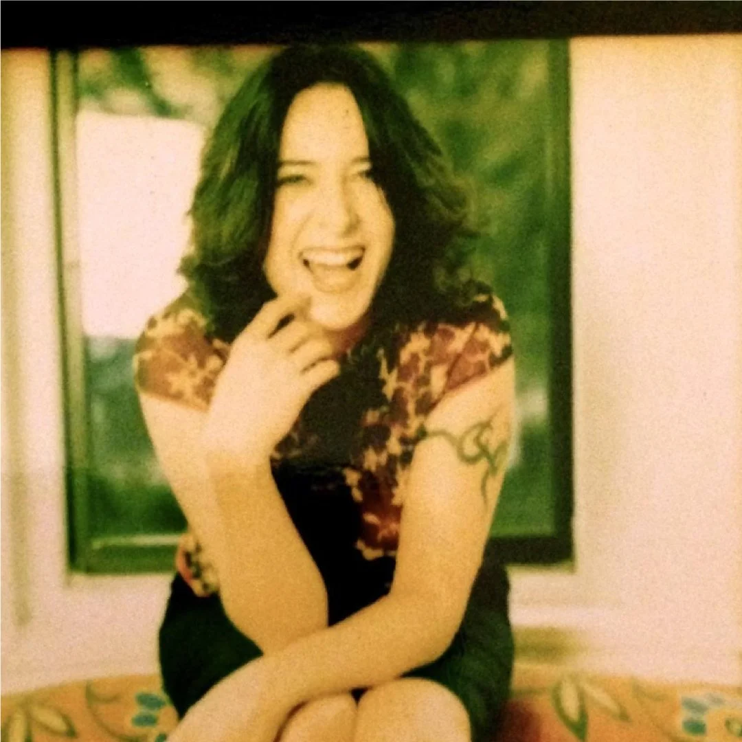 A woman with shoulder-length curly dark hair, smiling and winking, sitting indoors in front of a window with green scenery, wearing a colorful patterned top and black shorts.