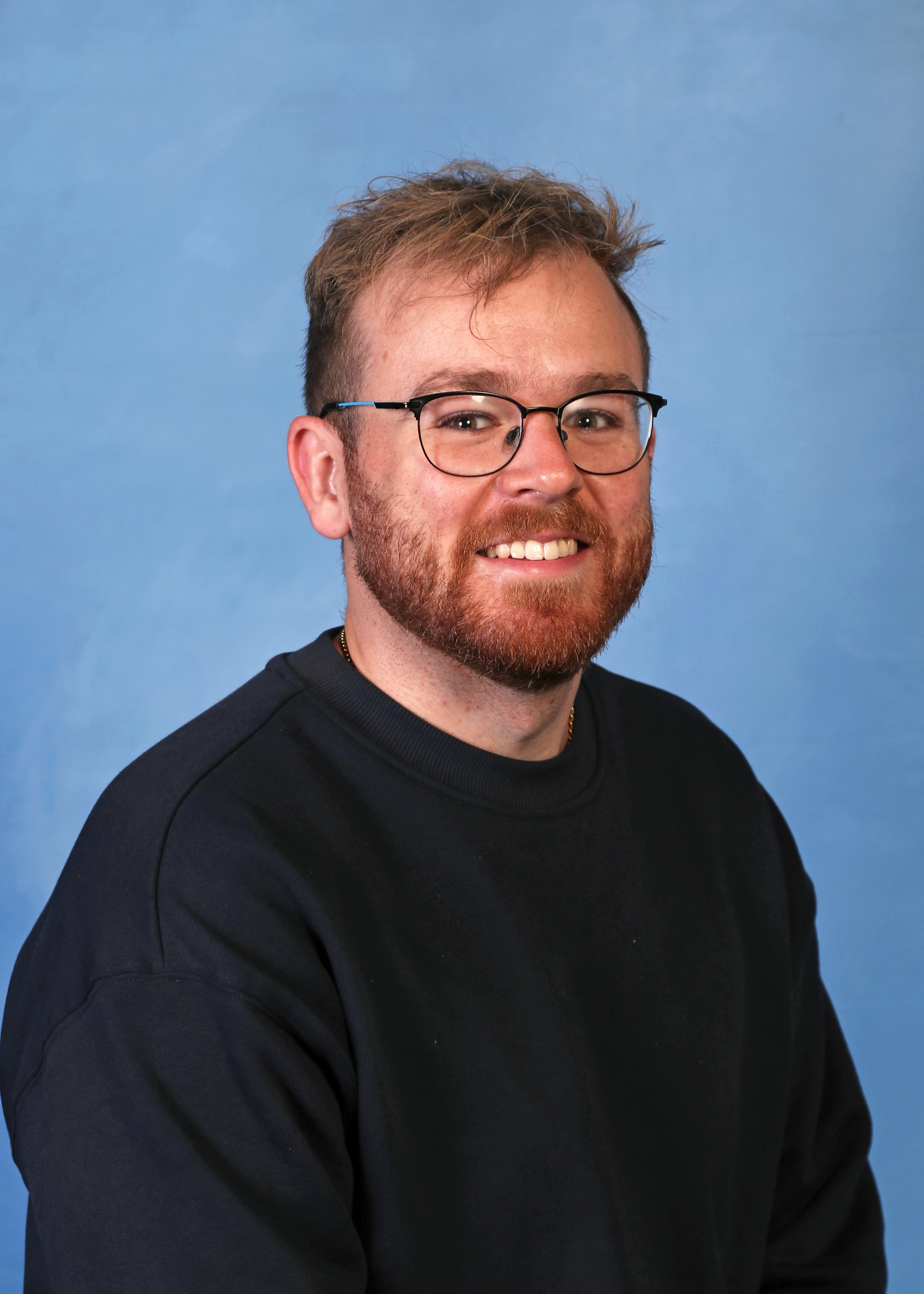 Headshot of a smiling man with glasses, a beard, and short brown hair, wearing a black sweater, against a blue background.