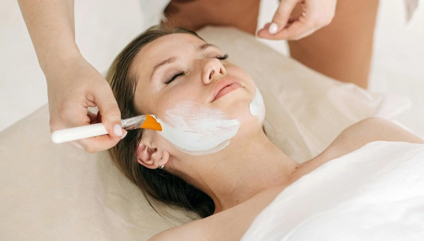 A woman receives a facial treatment with white cream applied to her cheeks and chin in a spa setting.