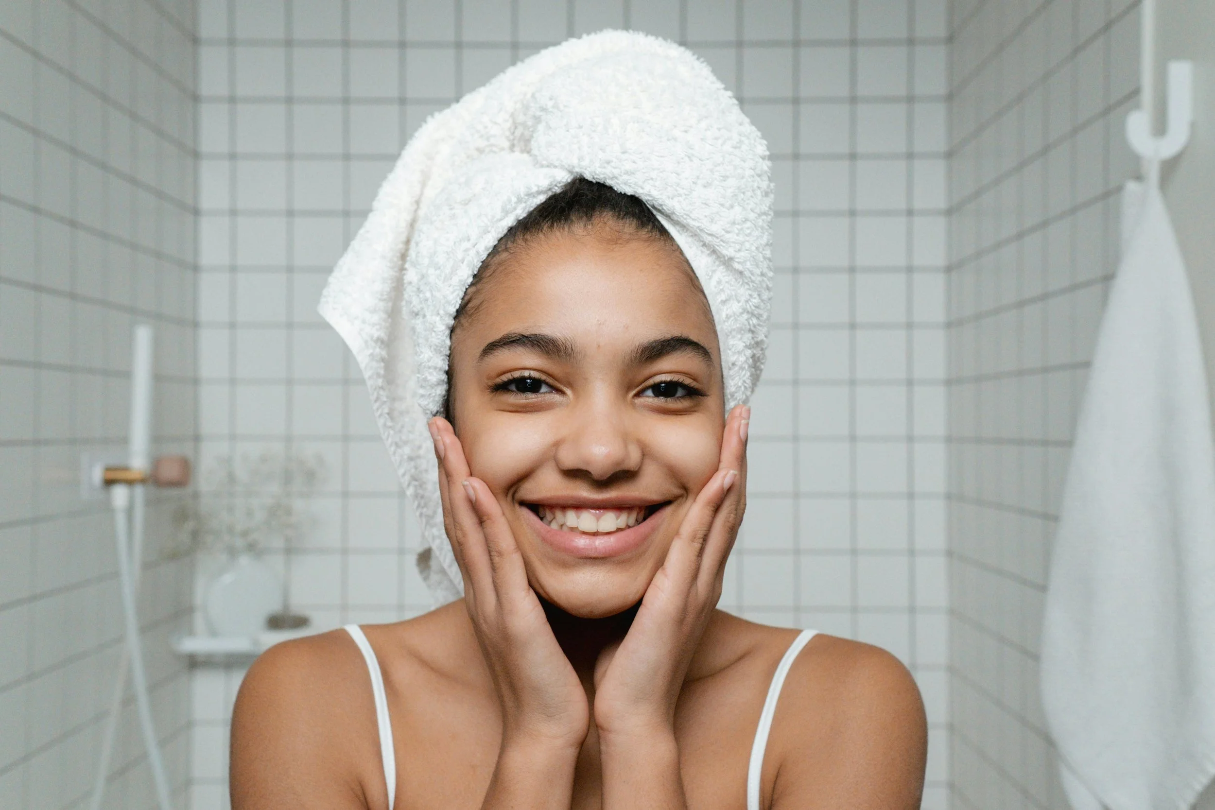 Young woman with a towel wrapped around her head, smiling and touching her cheeks in a bathroom with white tiled walls.