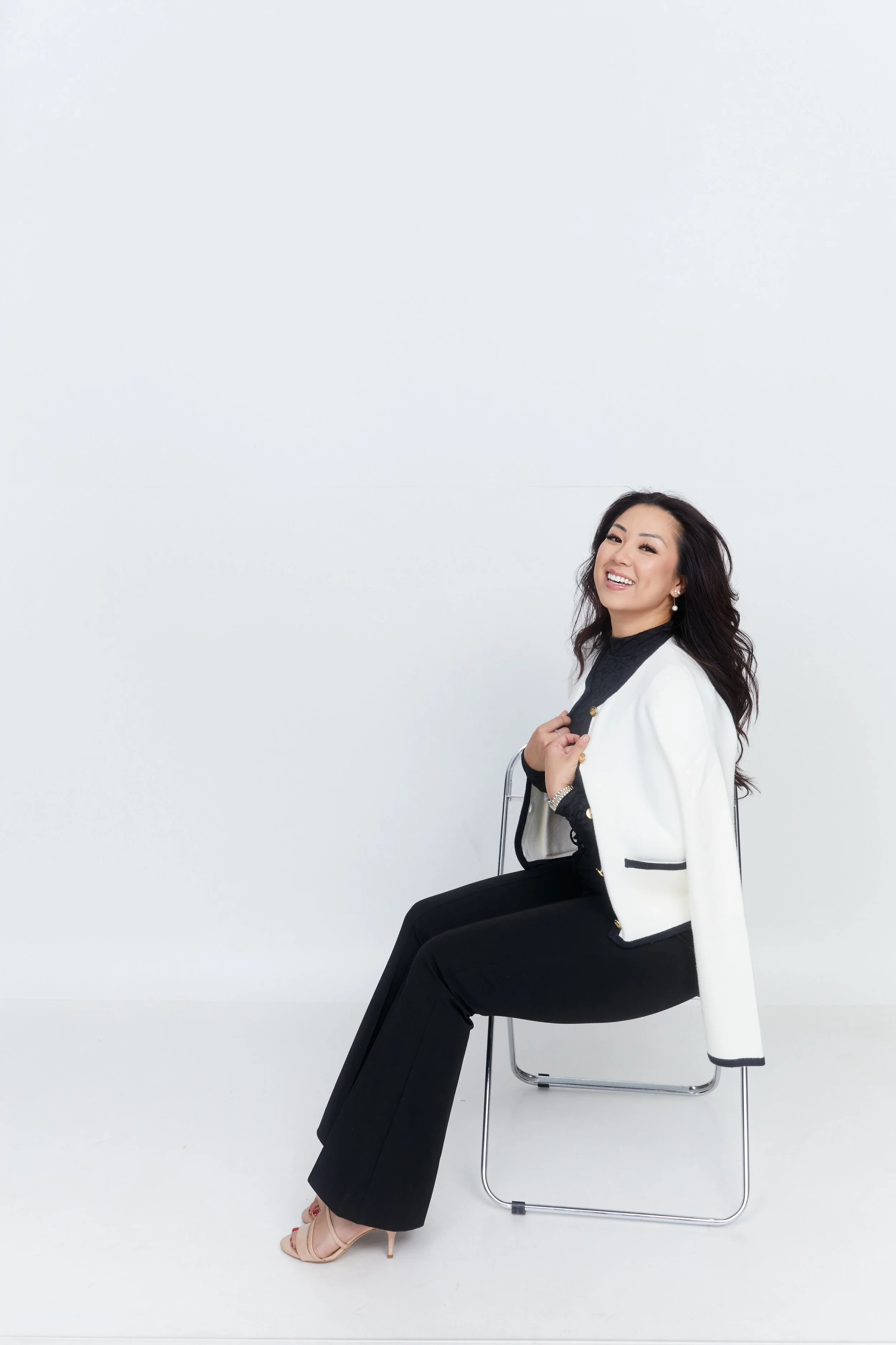 A woman with long wavy dark hair, wearing a white blazer, black top, black pants, and beige high heels, sitting on a metal chair against a plain white background, smiling and looking at the camera.