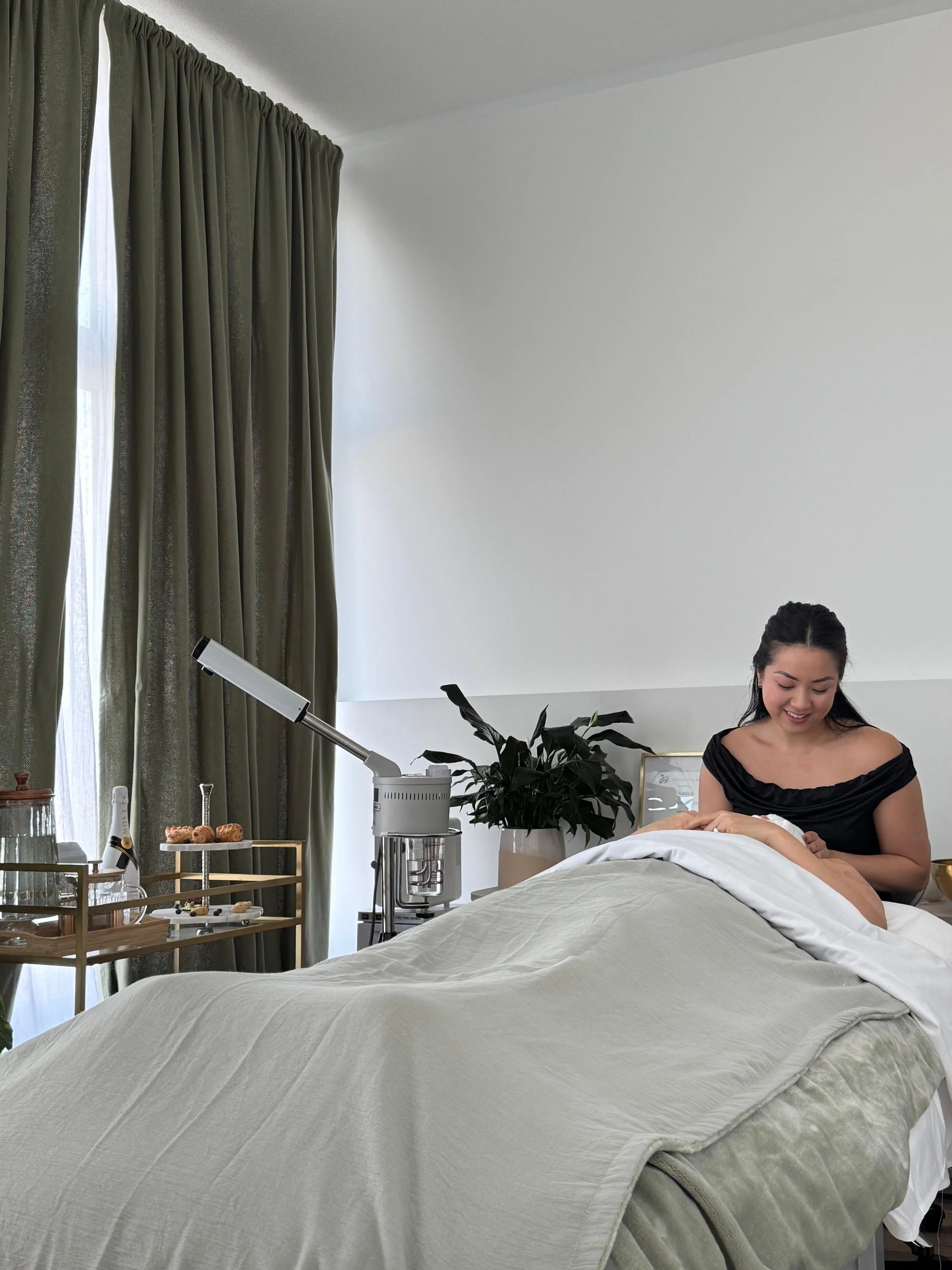 A woman smiling and preparing to give a massage in a well-lit room with green curtains, a large plant, a salon bed with a blanket, and a tray with skincare products.
