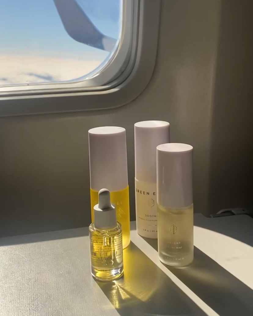 Skincare products on a tray on an airplane seat table, with sunlight and an airplane window in the background.