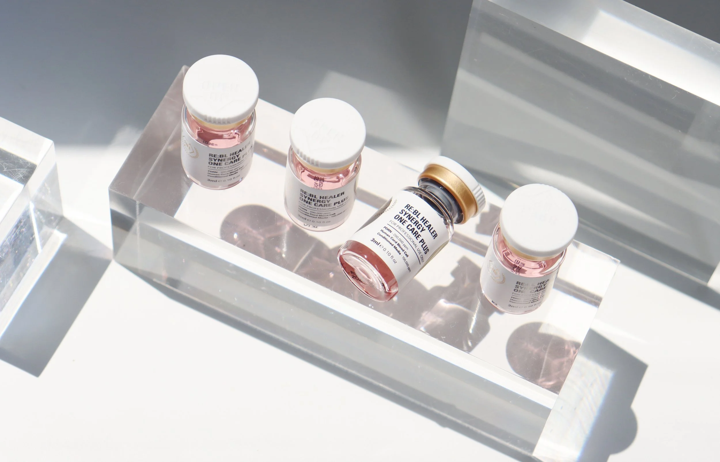 Four small bottles with pink liquid placed on a clear acrylic tray, with sunlight casting shadows.