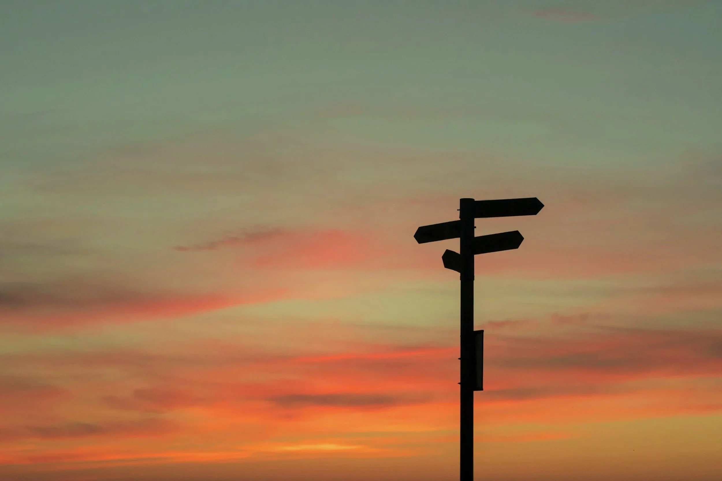 Silhouette of a street signpost against a colorful sunset sky with shades of pink, orange, and green.