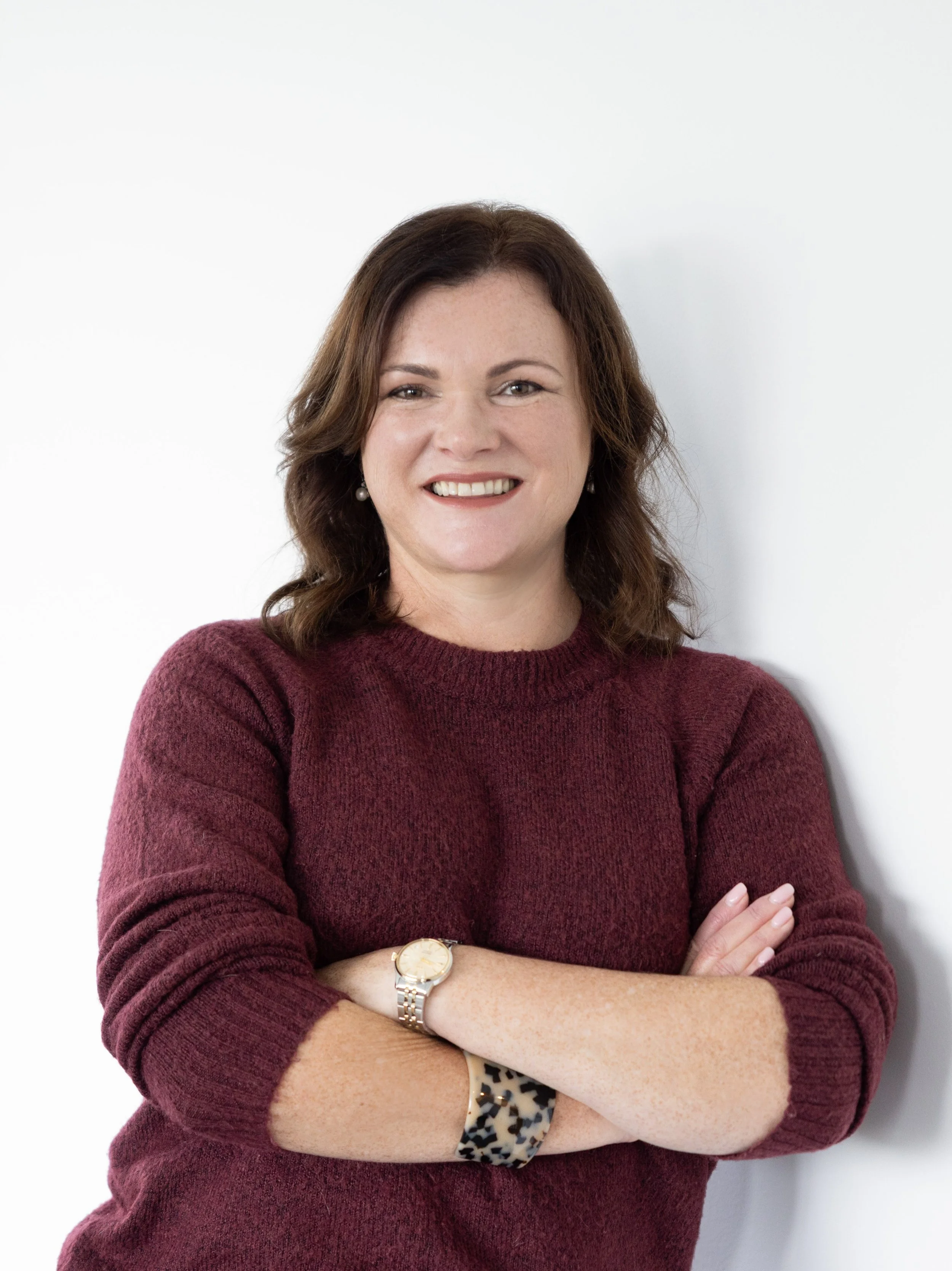 A woman with brown hair smiling, wearing a maroon sweater, crossing her arms, standing against a white background.