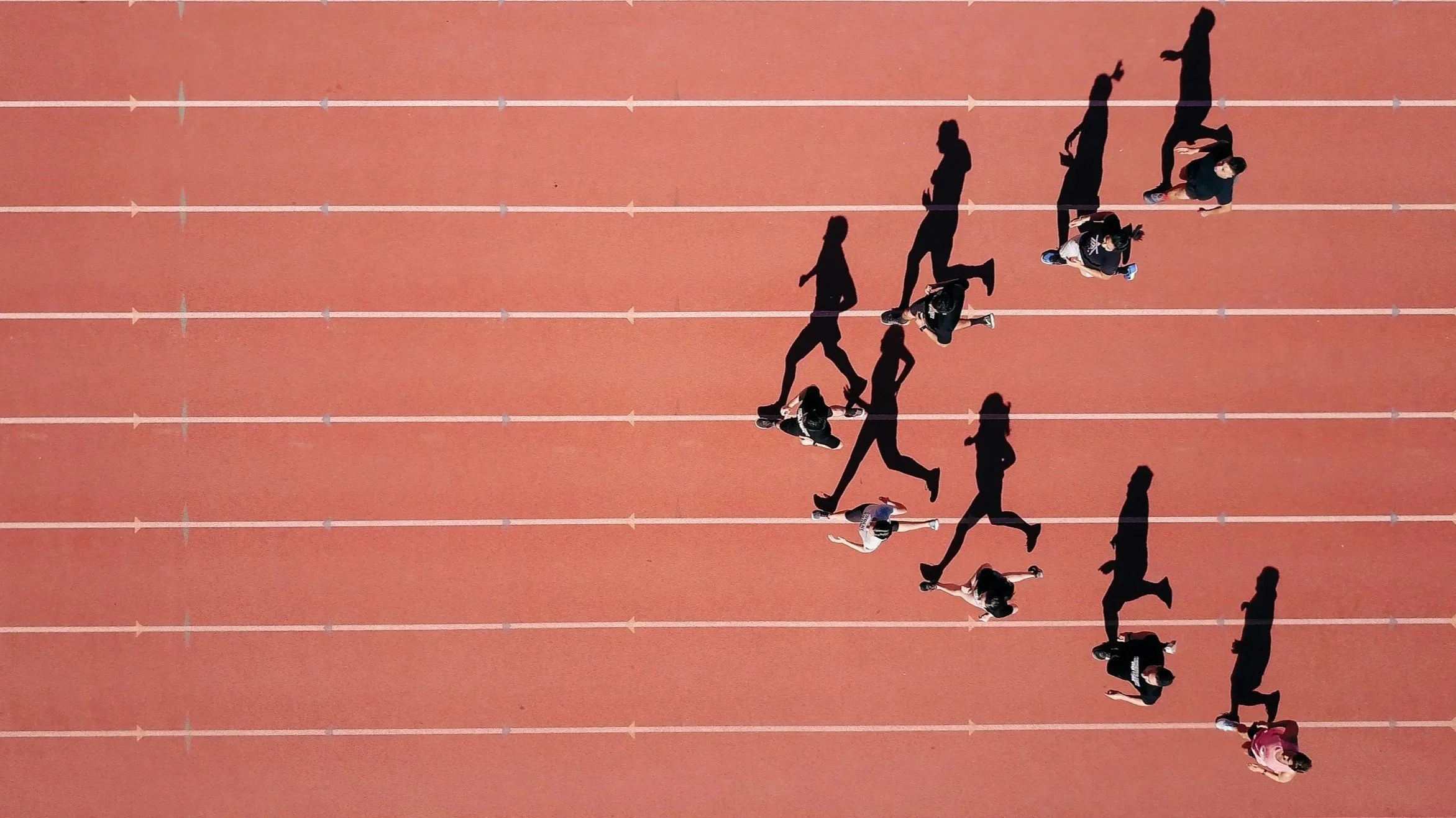 Top-down view of a red running track with multiple runners, some standing and some running, casting shadows on the track.