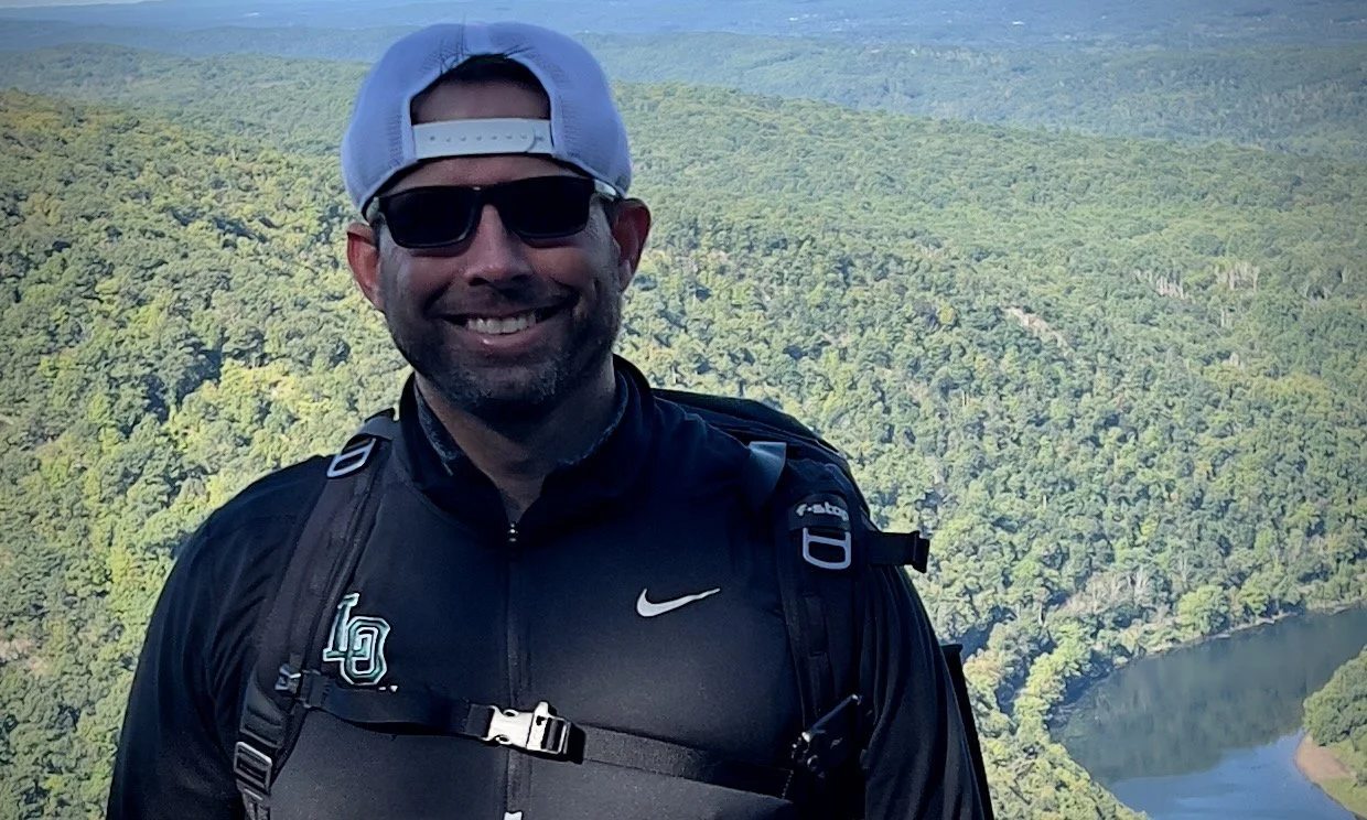Man smiling outdoors, wearing sunglasses, a black Nike sports jacket, a white cap backwards, and a backpack, with a forested landscape and a river below in the background.