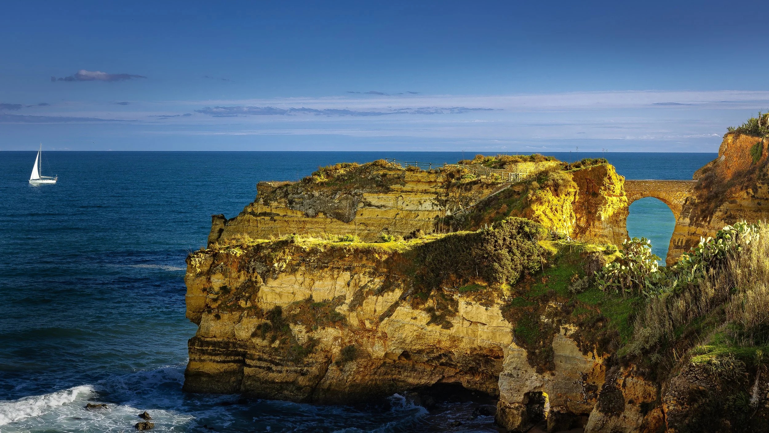 Cliffs by the ocean with a natural arch, a sailboat in the water, and a cloudy sky.