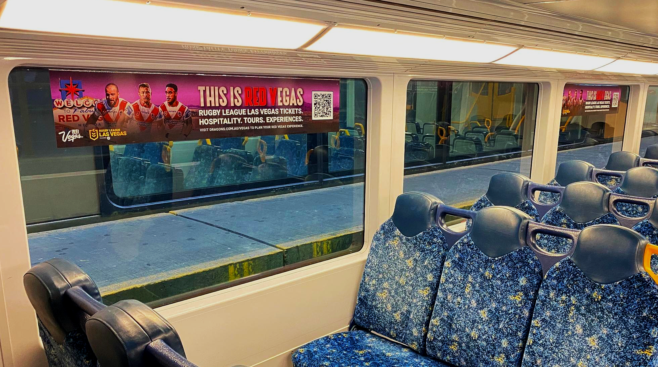 Inside a train, with blue patterned seats and a large window showing an advertisement for Las Vegas, featuring rugby players and bold red and yellow text.