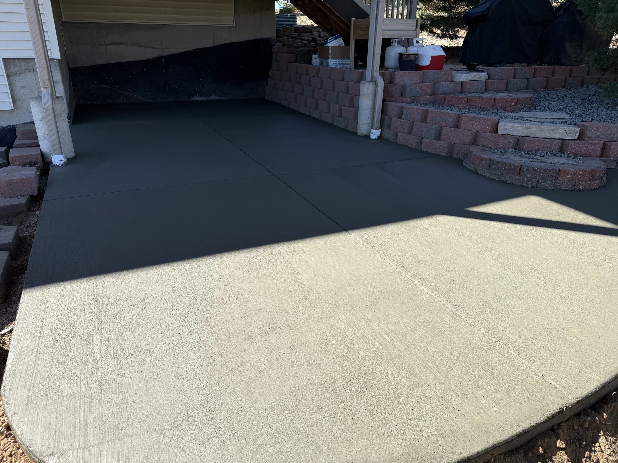 Freshly poured concrete on a driveway with a curved edge, adjacent to a house with a brick retaining wall and gardening supplies.
