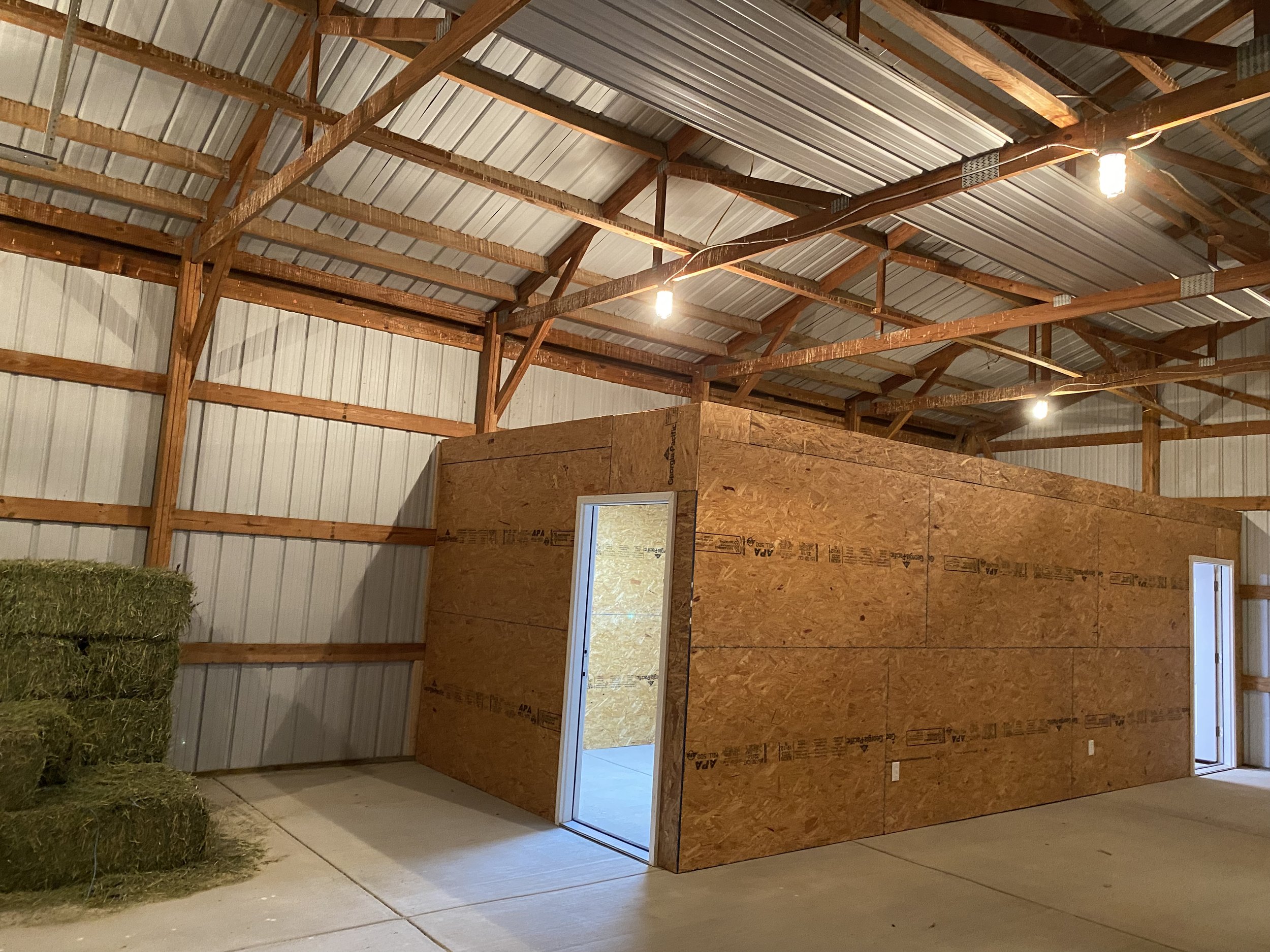 An unfinished wooden structure inside a barn or shed, with walls made of oriented strand board (OSB), a metal roof supported by wooden beams, and a concrete floor. There is a doorway and a window opening in the wall, and a stack of hay bales is visible on the left side.