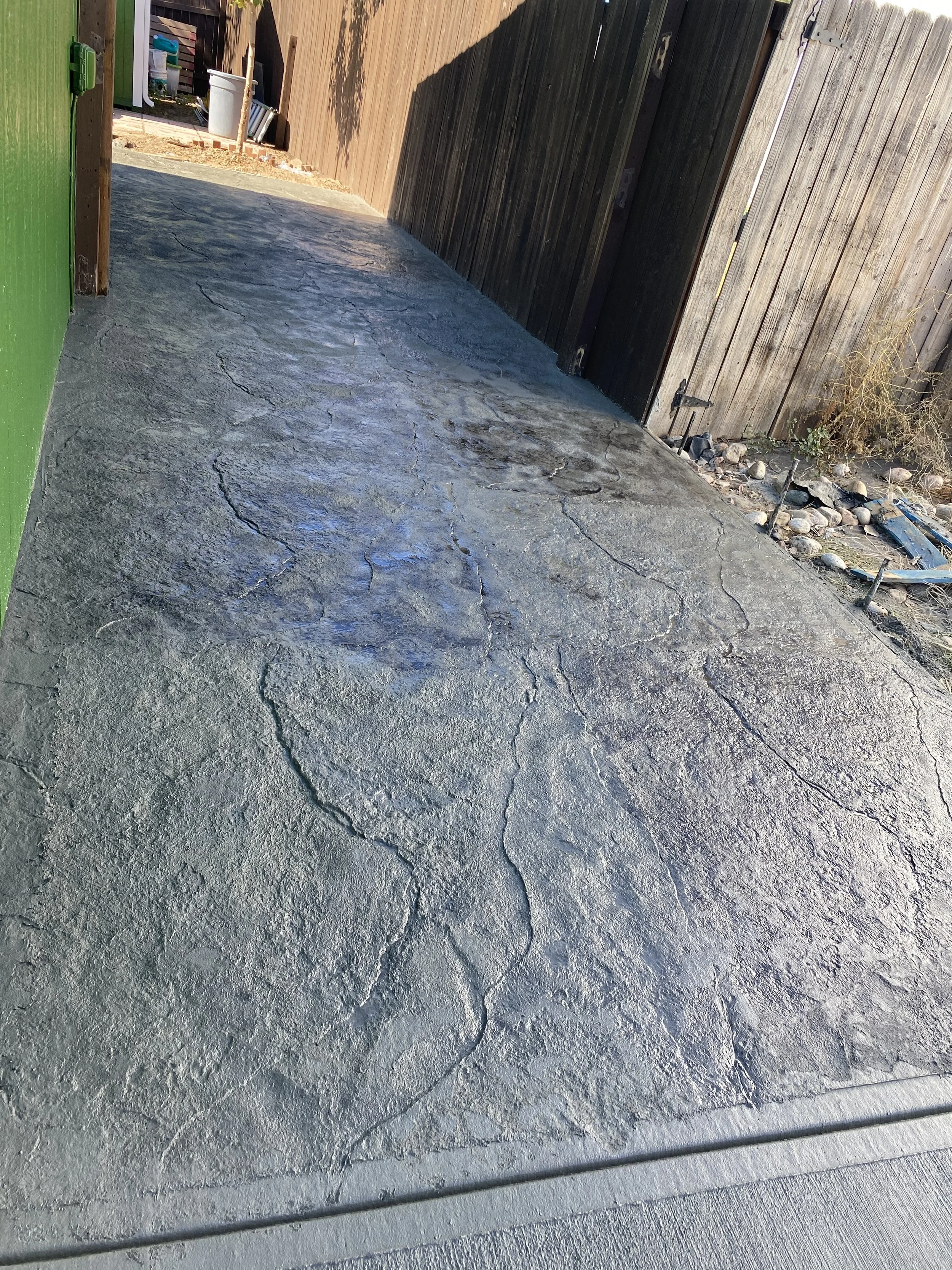 A concrete backyard patio with cracks, bordered by a green wall on the left and a wooden fence on the right, with a garden bed and rocks in the background.