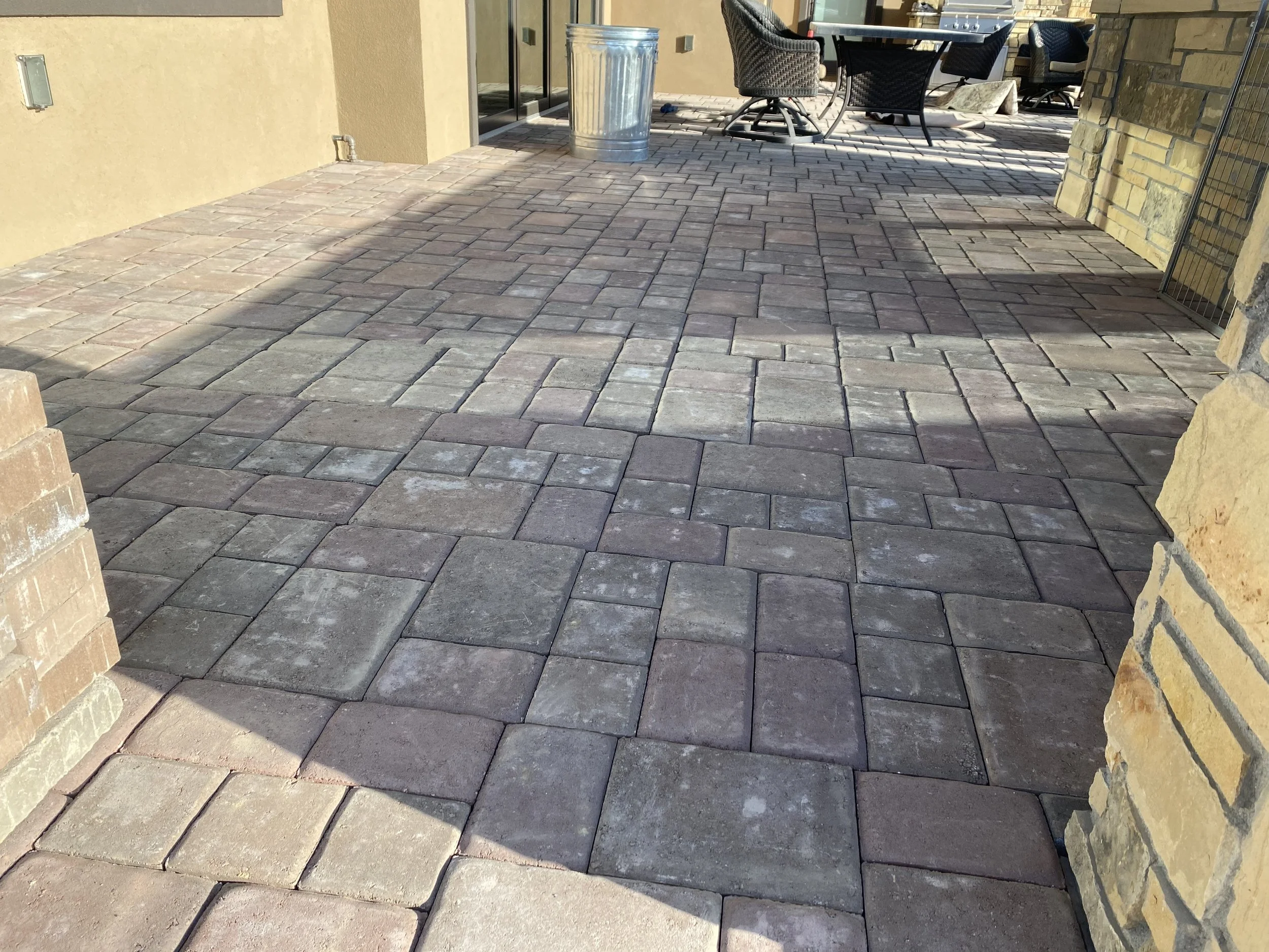 Paved outdoor patio with stone bricks, glass table, chairs, and a trash can in the background.