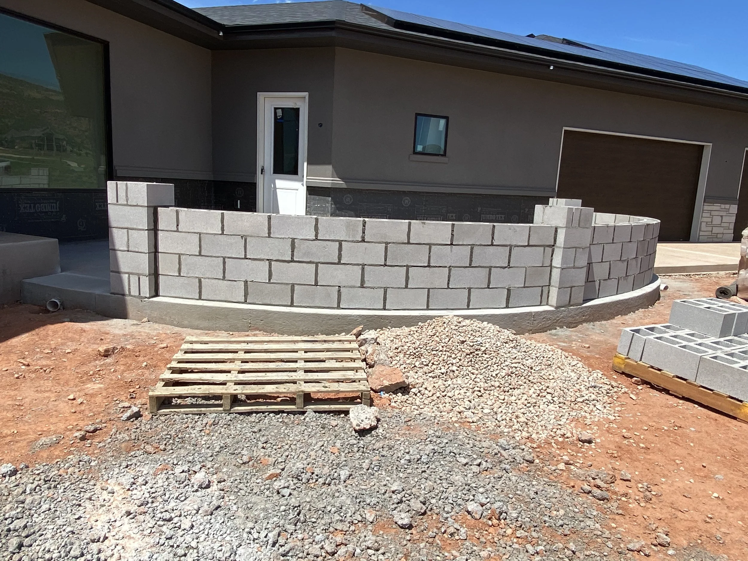 Construction site with a curved cinder block wall building feature in front of a house, with piles of gravel, a wooden pallet, and cinder blocks on the ground.
