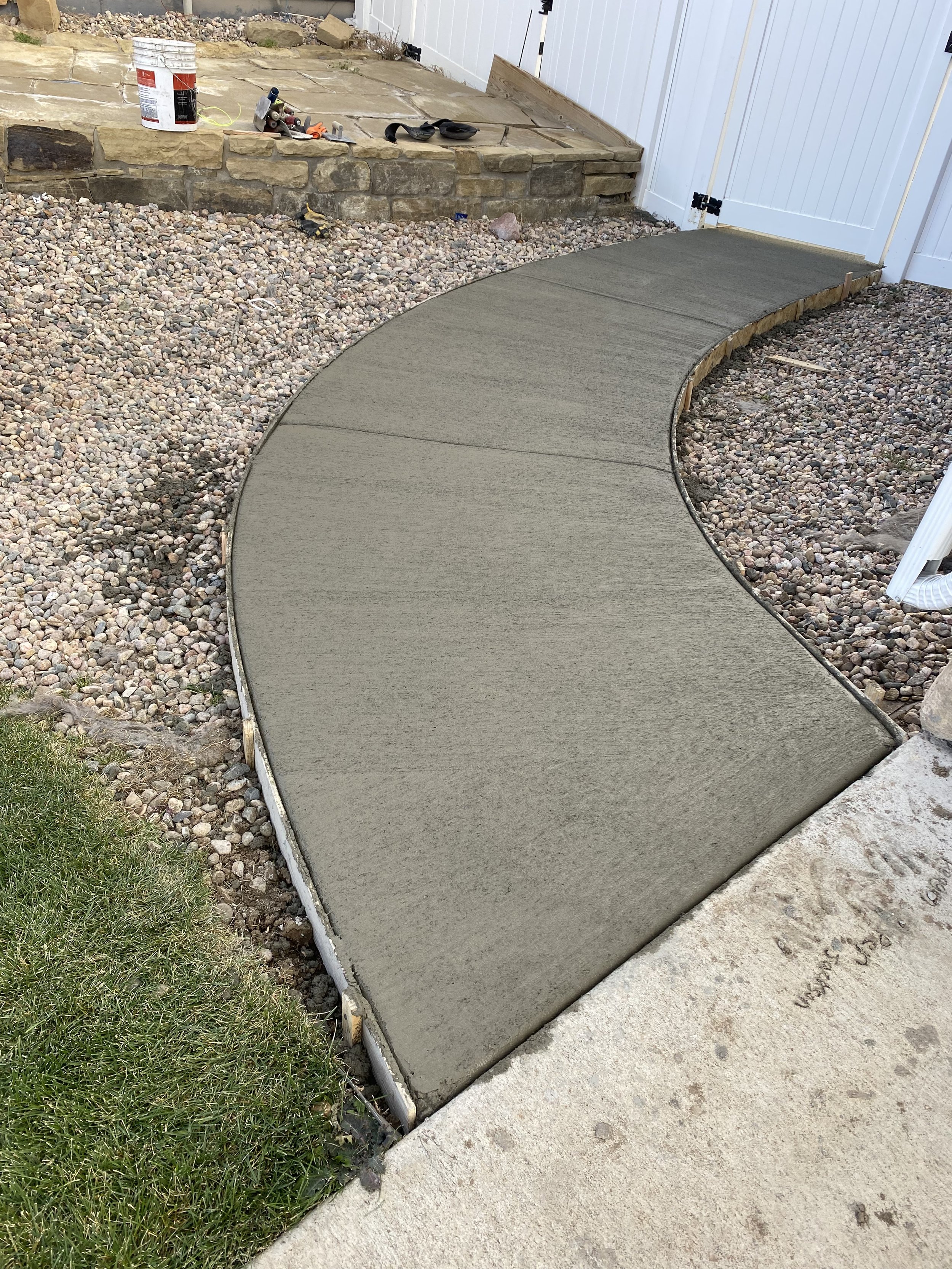 Recently poured concrete sidewalk with a curved shape alongside a gravel border and a grass patch, leading to a white fence and a closed white gate.