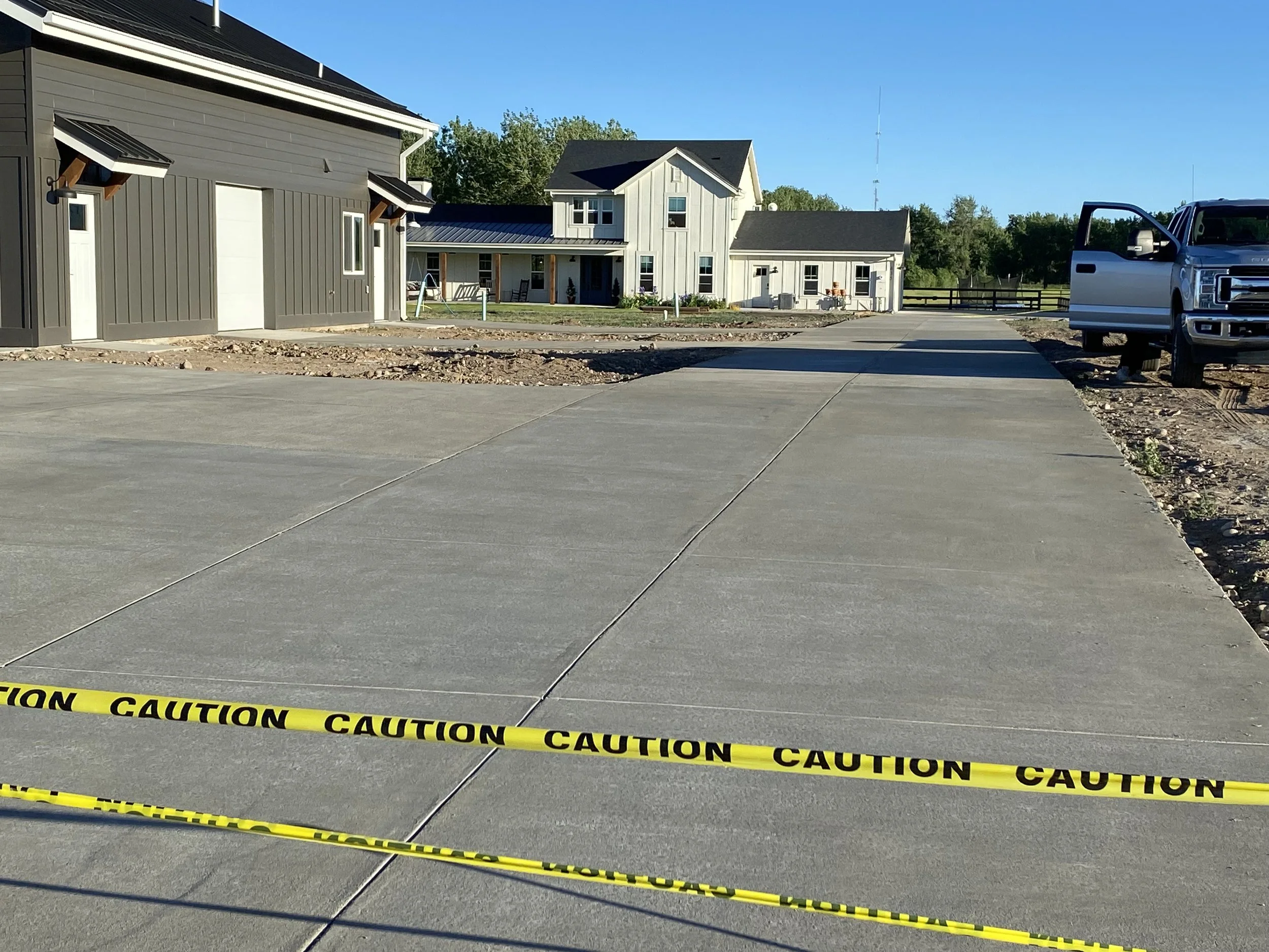Construction site with newly paved concrete driveway blocked off by yellow caution tape, houses in the background, and a pickup truck on the right side.