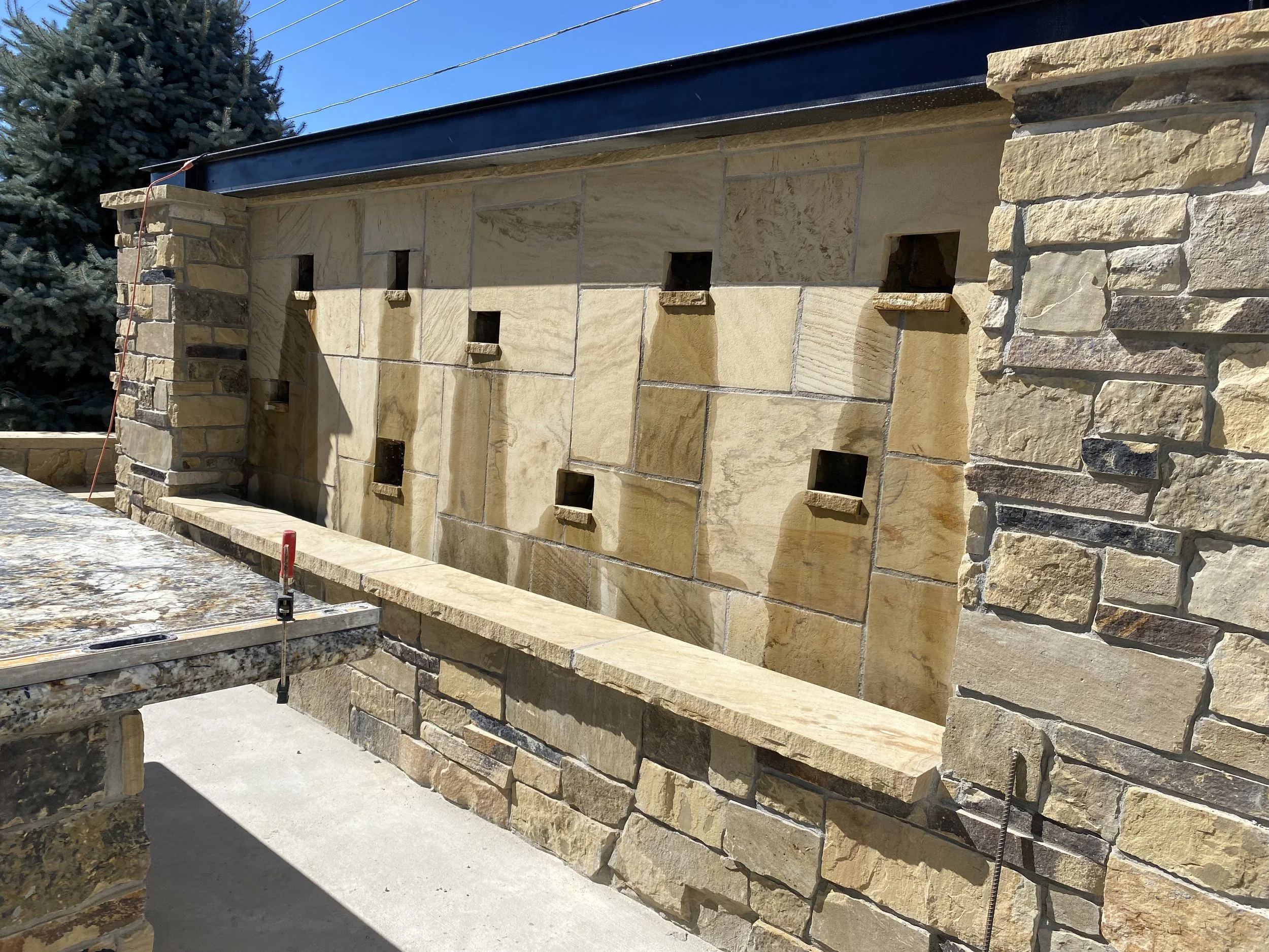 Close-up of a stone fountain wall under construction, with multiple small square openings and a ledge, showing beige and tan stone tiles and bricks, with some black metal elements and construction tools.