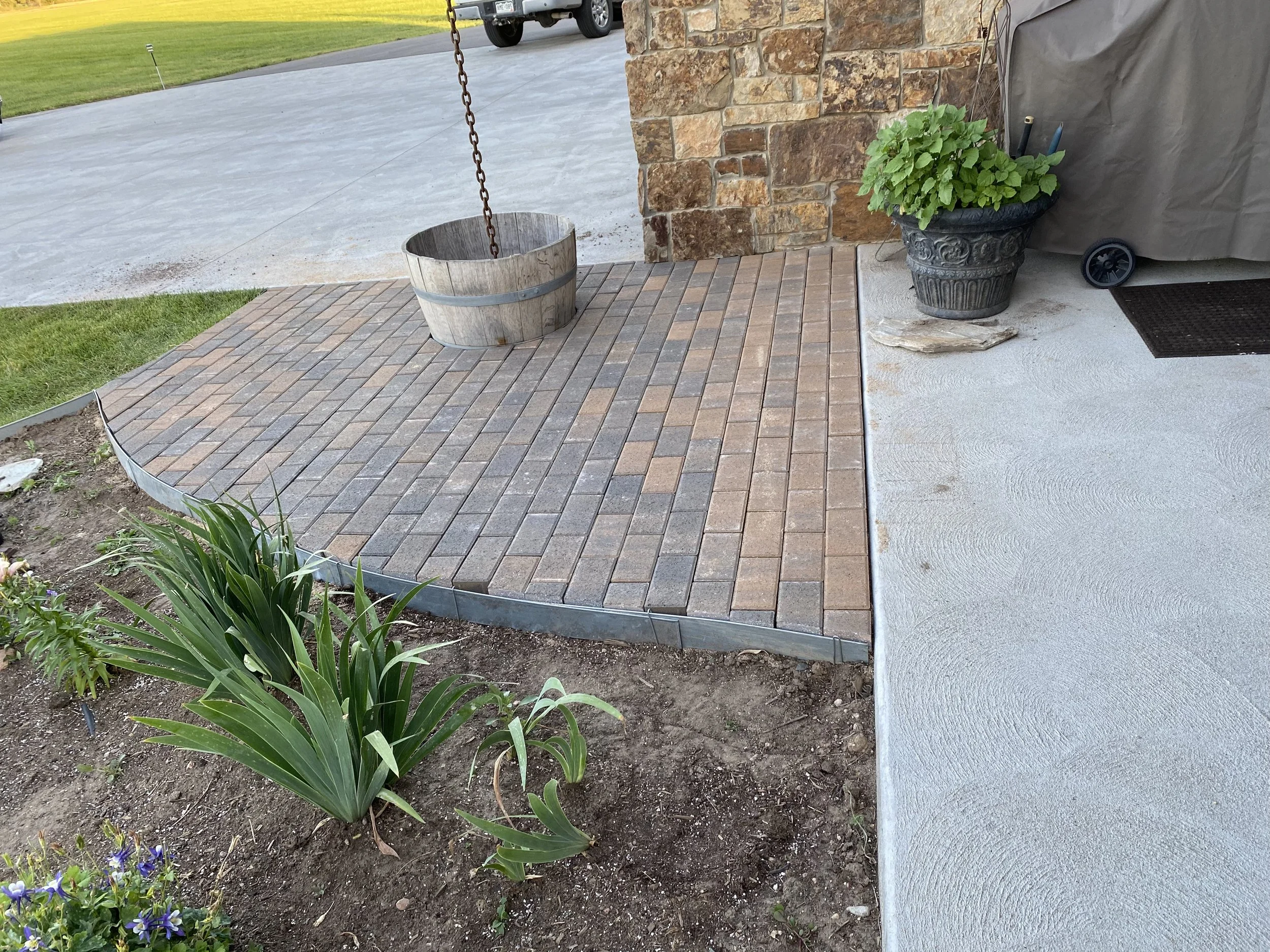 A small brick patio area next to a house with a stone wall. There is a hanging bucket, a large potted plant, and a covered grill. In the foreground, there are green plants and flowers in a garden bed.