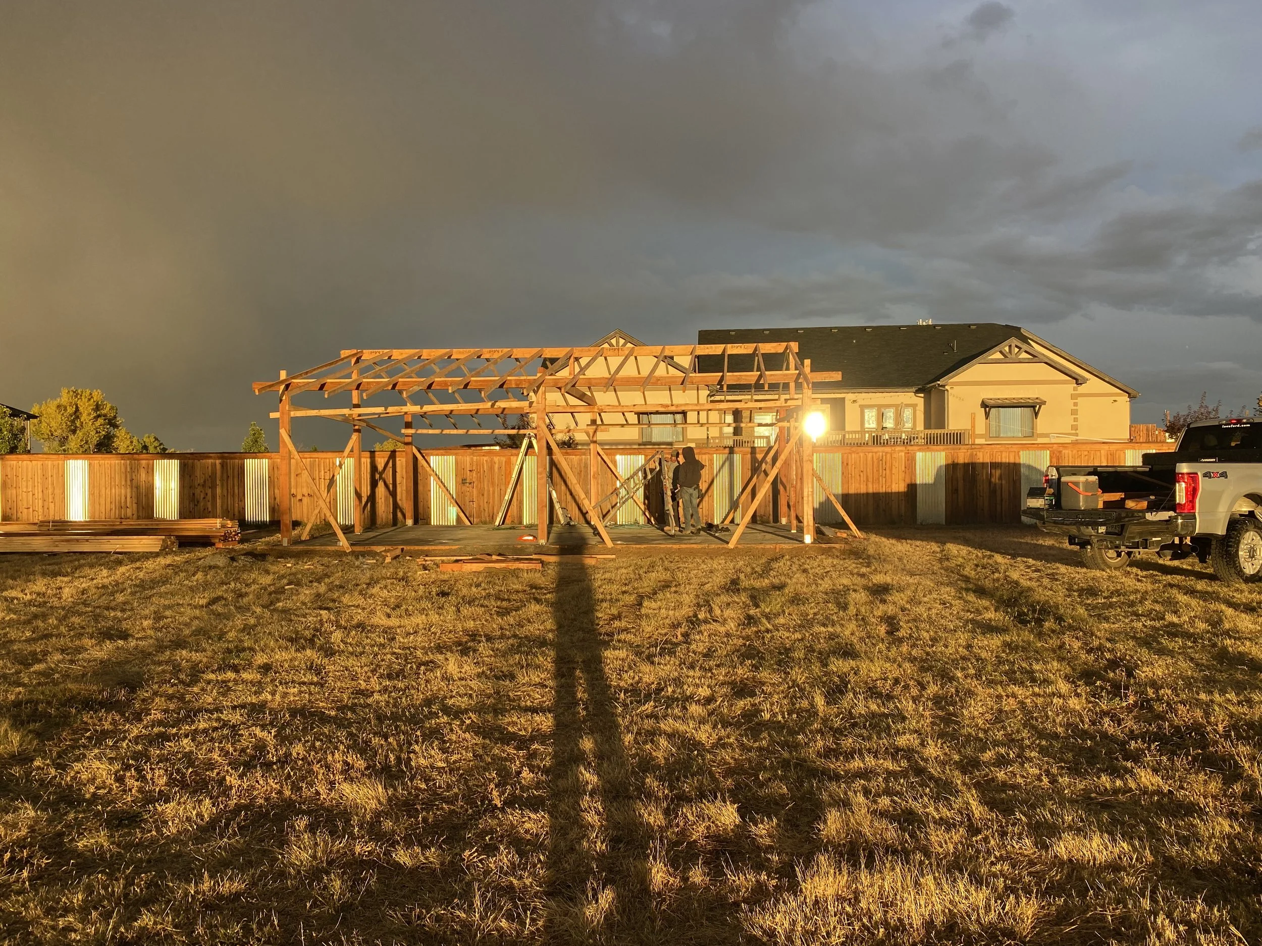 Construction of a wooden backyard patio or pergola with workers, during sunset, in a suburban backyard with a house and trucks nearby.
