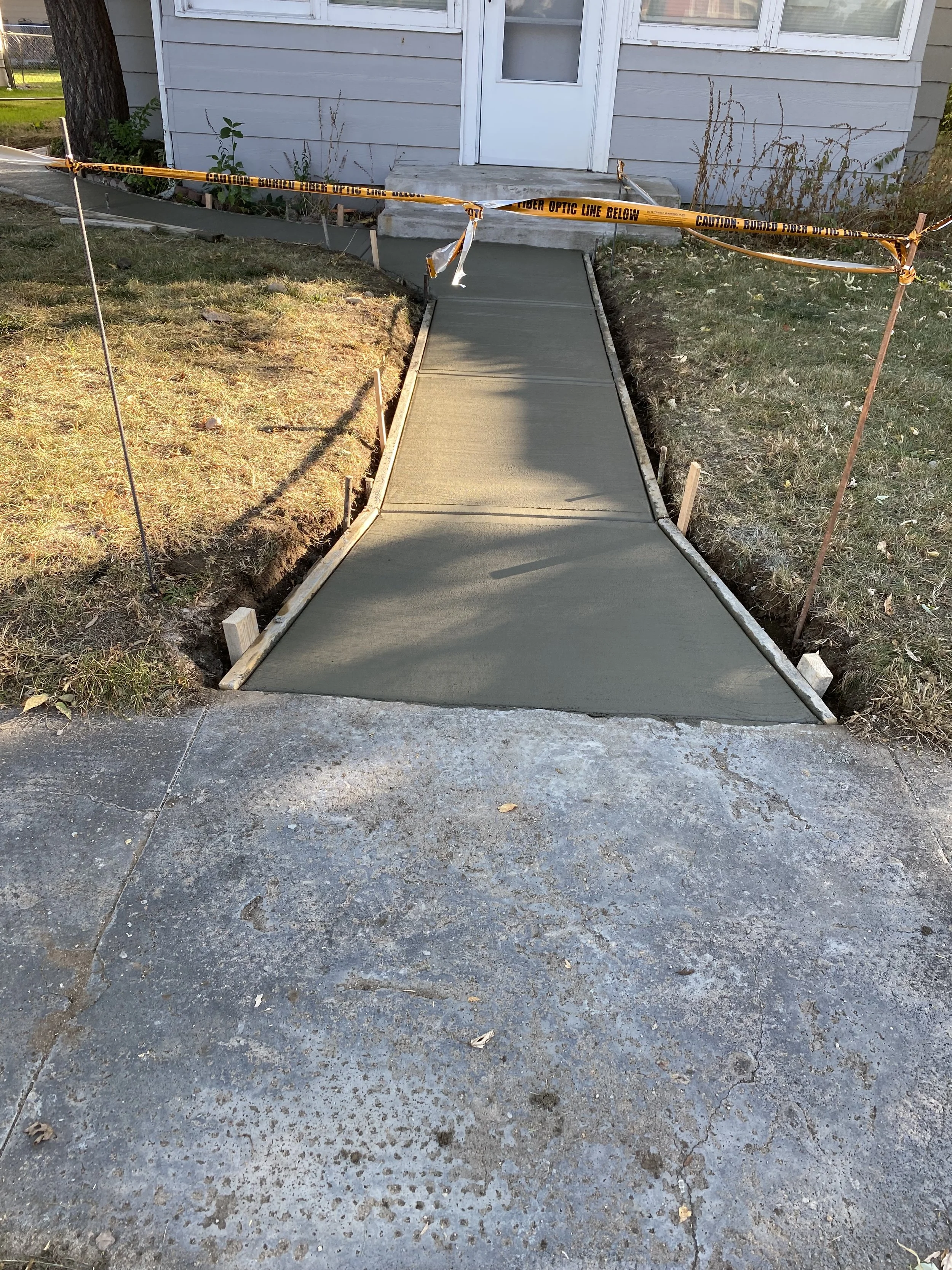 Newly poured concrete sidewalk leading to the front door of a house, surrounded by caution tape and construction stakes.