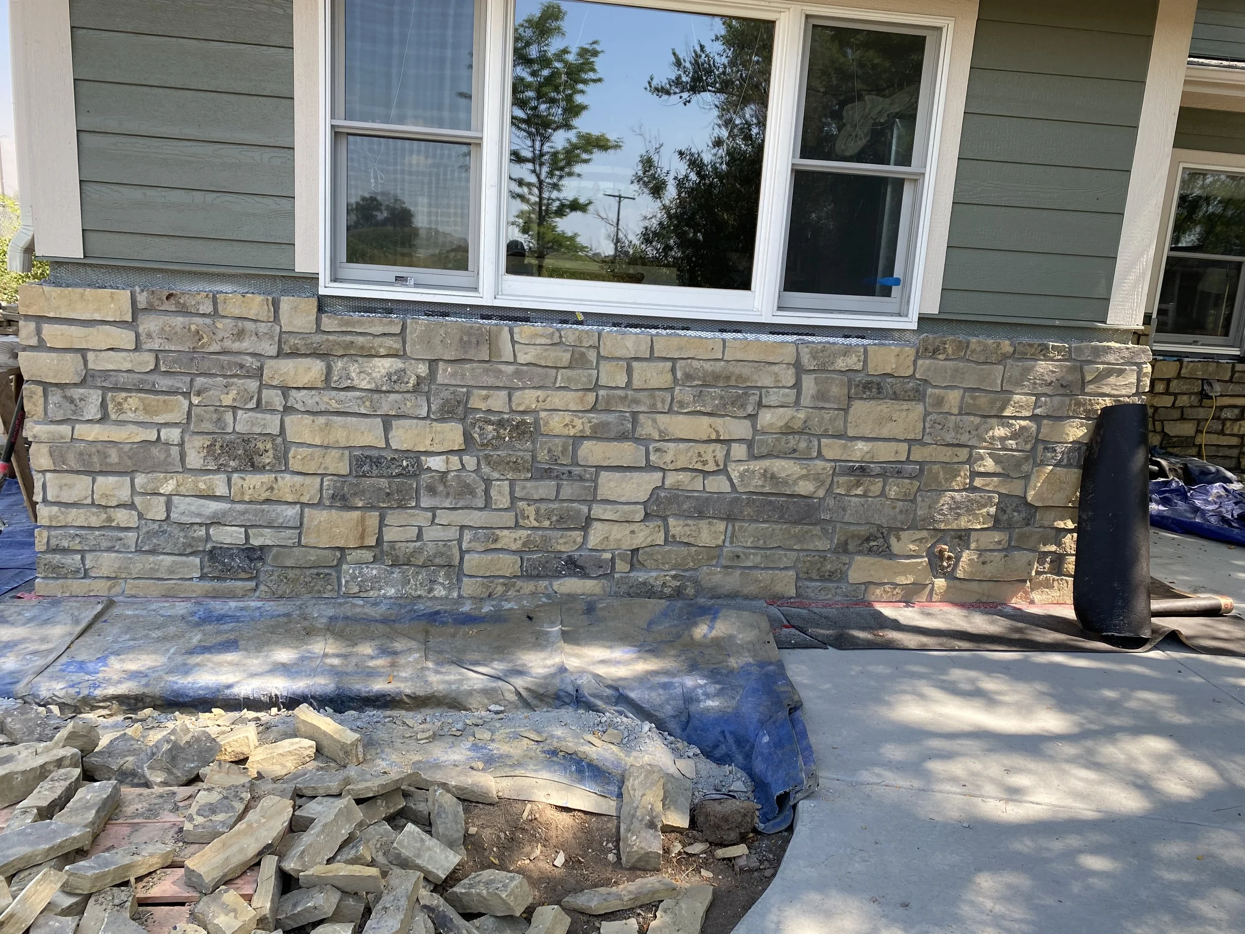 Construction site with a stone wall partially built under a window of a house, with construction materials and tools scattered around.
