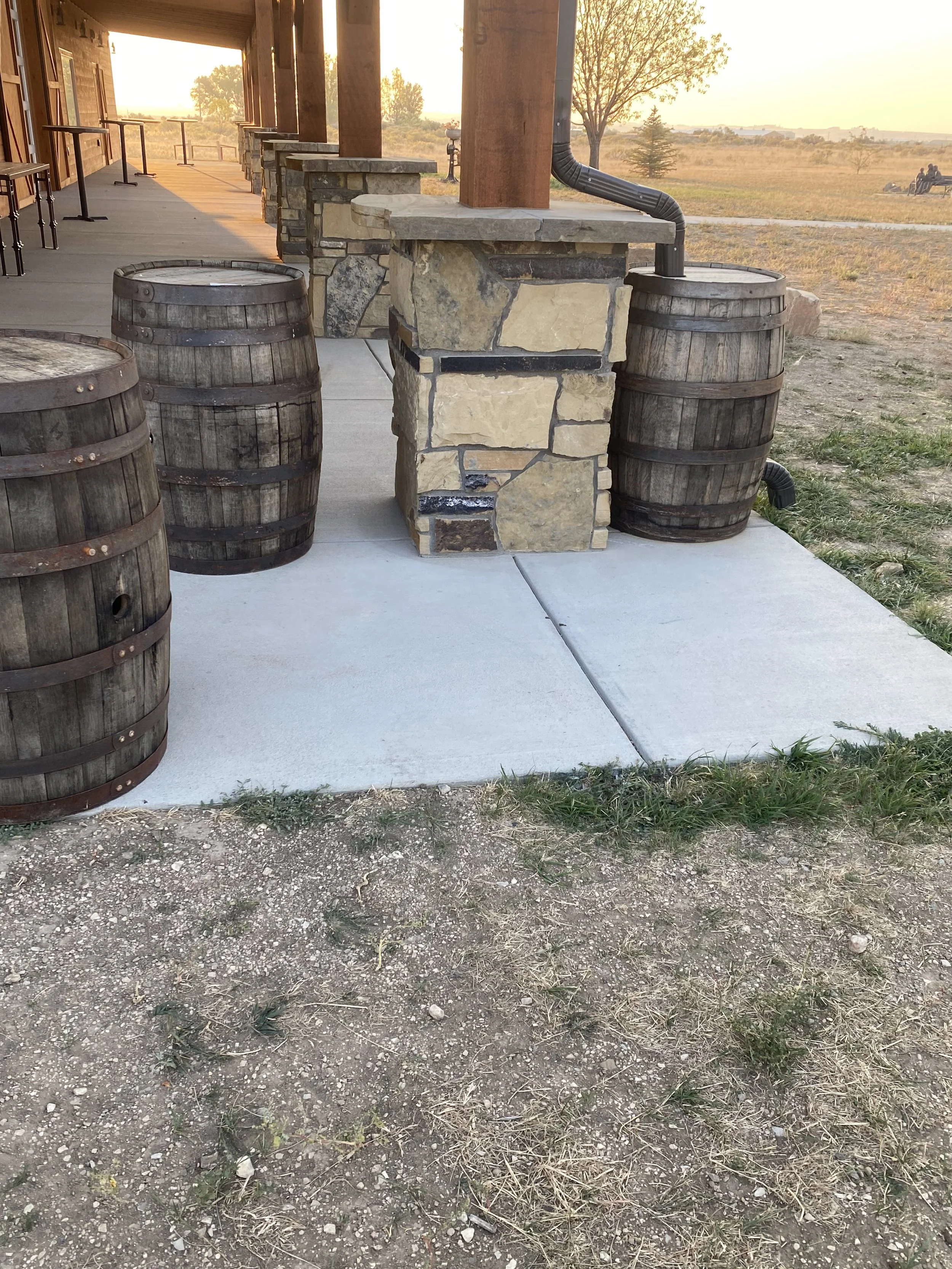 A rustic outdoor patio area with a stone and wood bar counter, surrounded by three wooden barrel seats on a concrete slab, with a rural landscape and trees in the background at sunset.