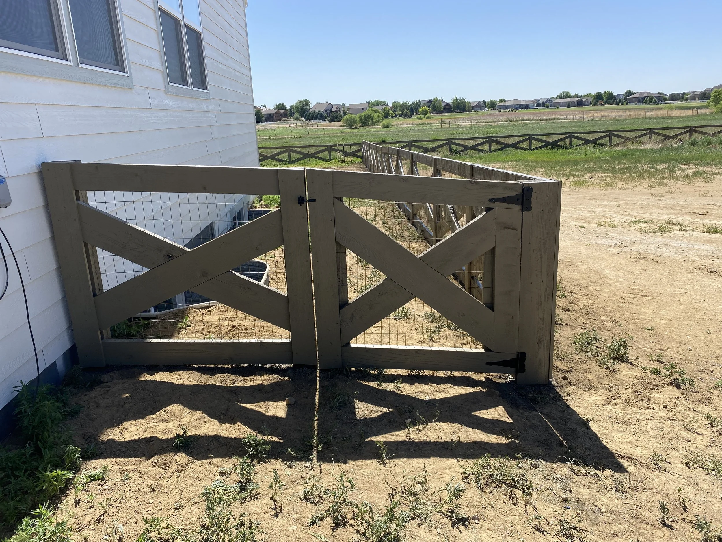 Wooden gate with wire mesh at the side of a white house, leading to an open field with grass and distant houses.