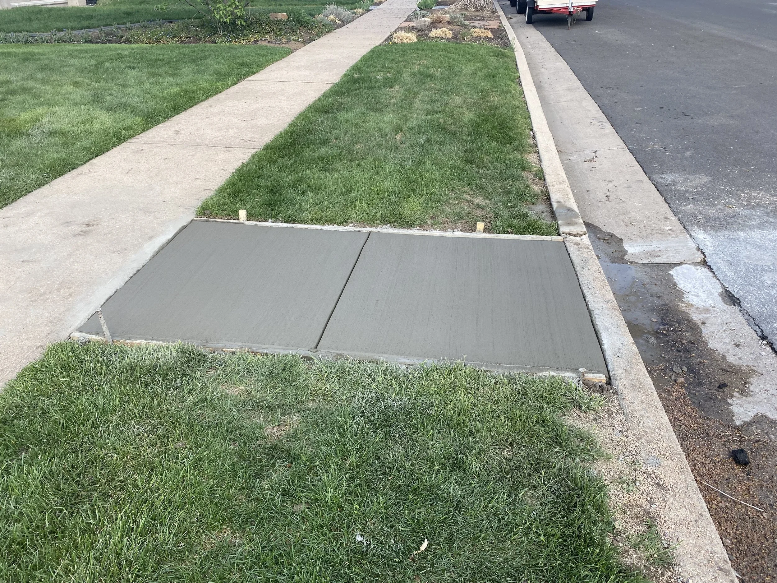Freshly poured concrete sidewalk section adjacent to grass and a curb, with a town street and a parked trailer visible in the background.