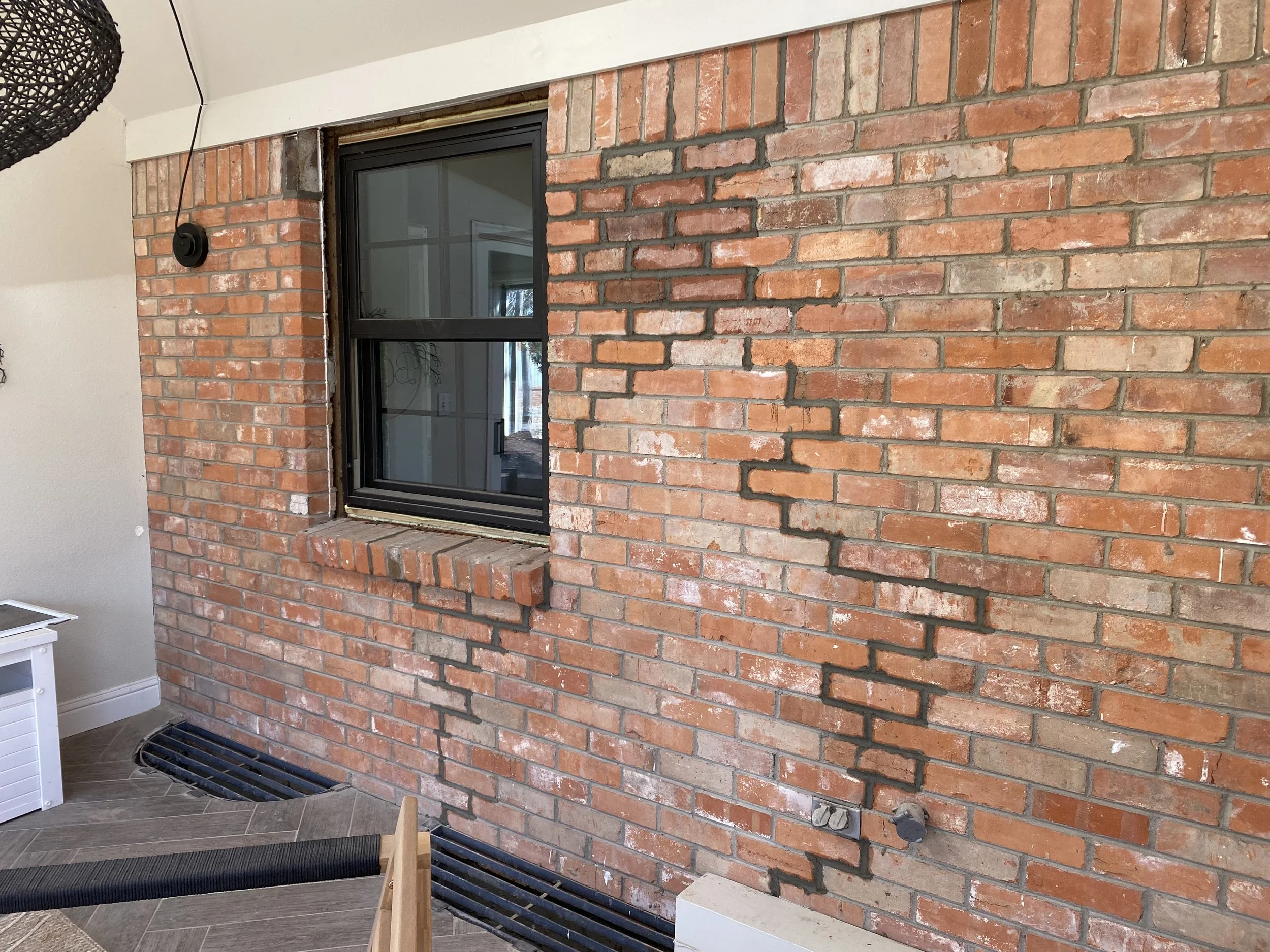 Interior brick wall with window and electrical outlets, with black air vents on the floor.
